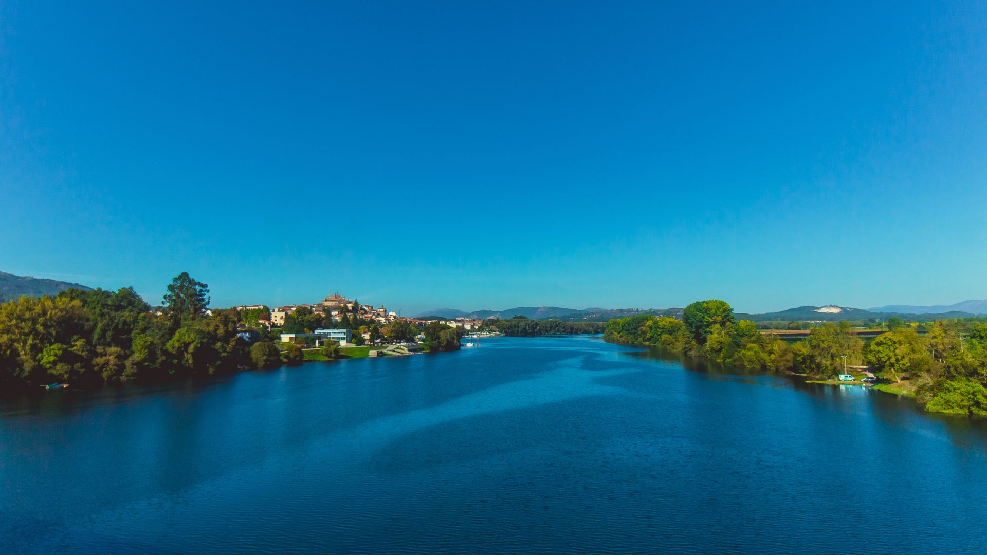 A panoramic view of the Minho River with the historic town of Valença do Minho on its bank, surrounded by lush green hills under a clear blue sky.