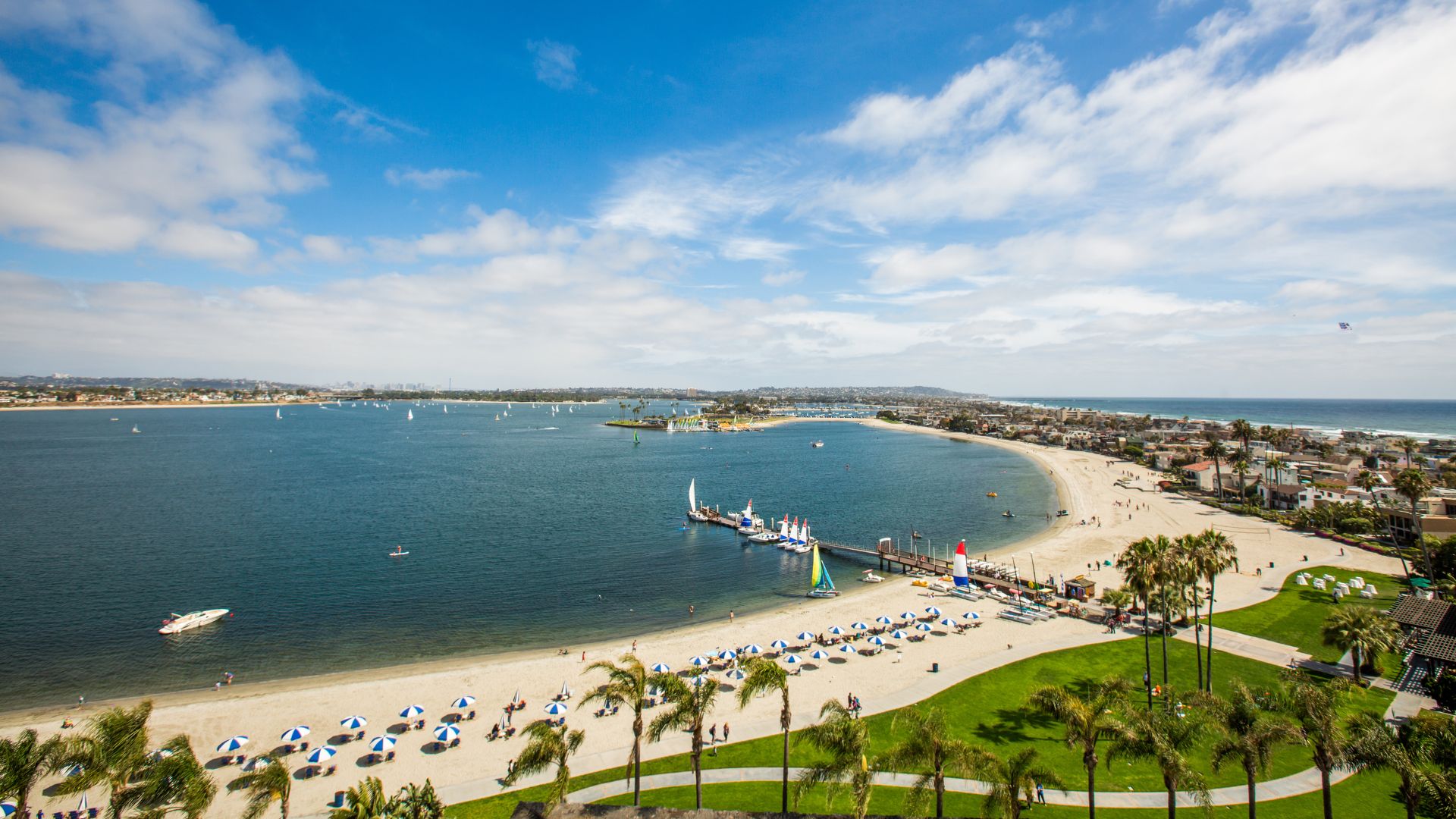 Aerial view of Mission Beach in San Diego, California, showcasing the narrow strip of land with houses and a road, bordered by the Pacific Ocean on one side and Mission Bay with boats and a distant city skyline on the other.