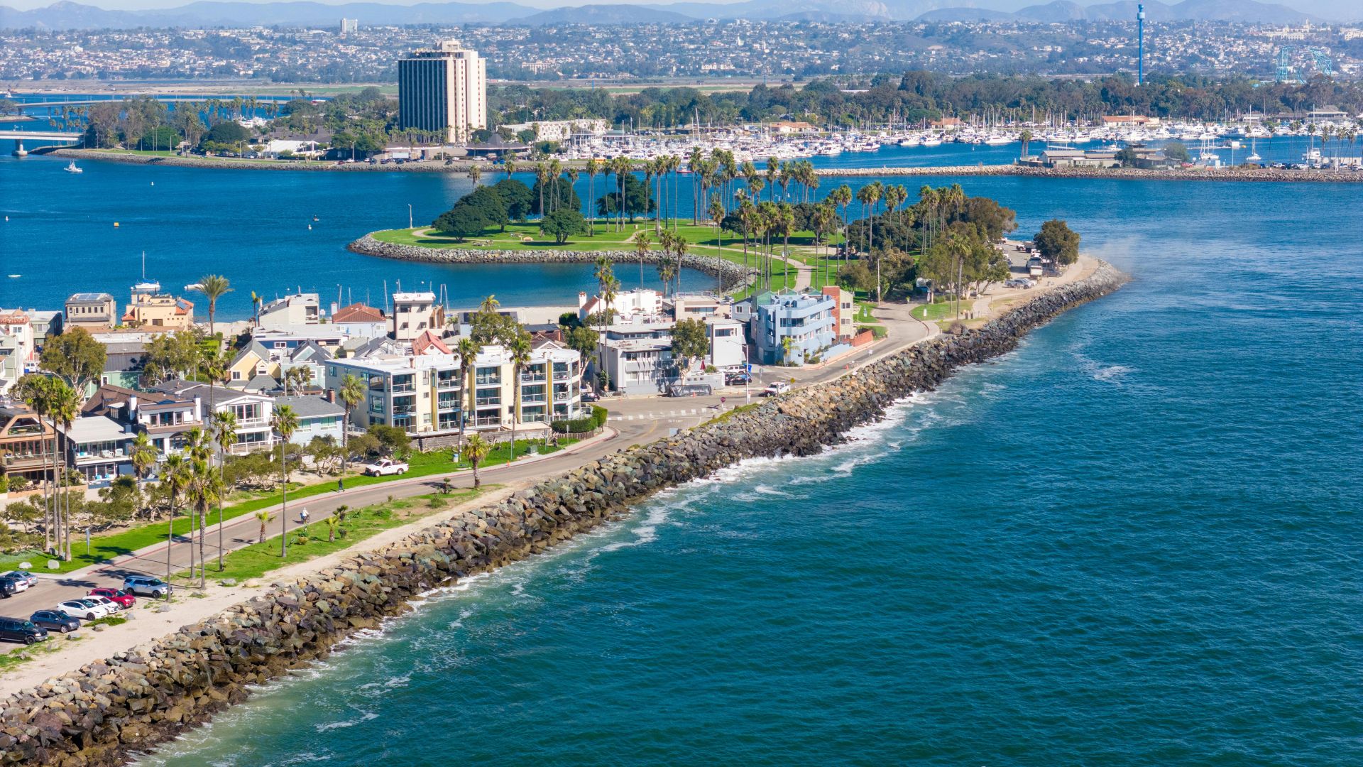 Aerial view of Mission Beach in San Diego, California, showcasing the narrow strip of land with houses and a road, bordered by the Pacific Ocean on one side and Mission Bay with boats and a distant city skyline on the other.