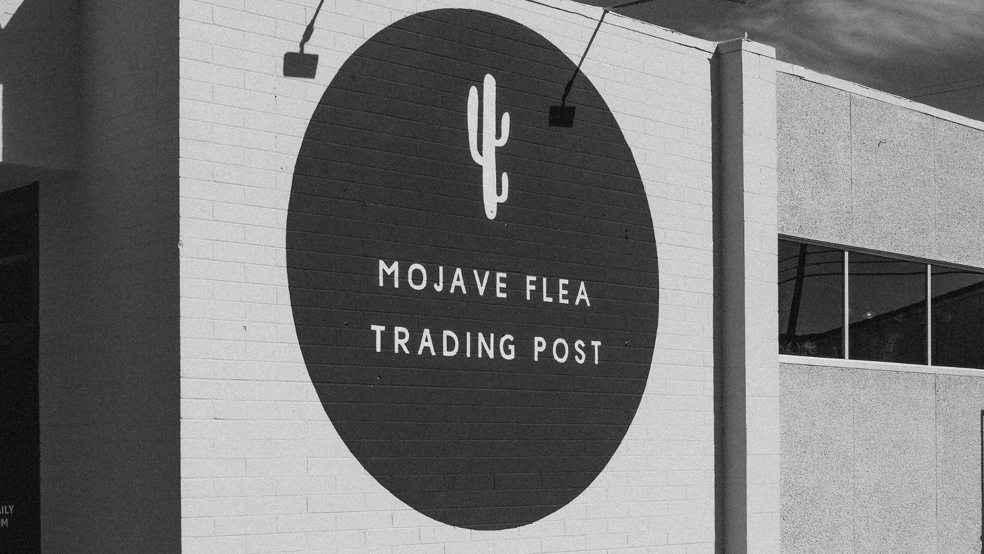 A black and white photo of the exterior of a building with "MOJAVE FLEA TRADING POST" and a cactus logo painted in white on a large black circle on its white brick wall.