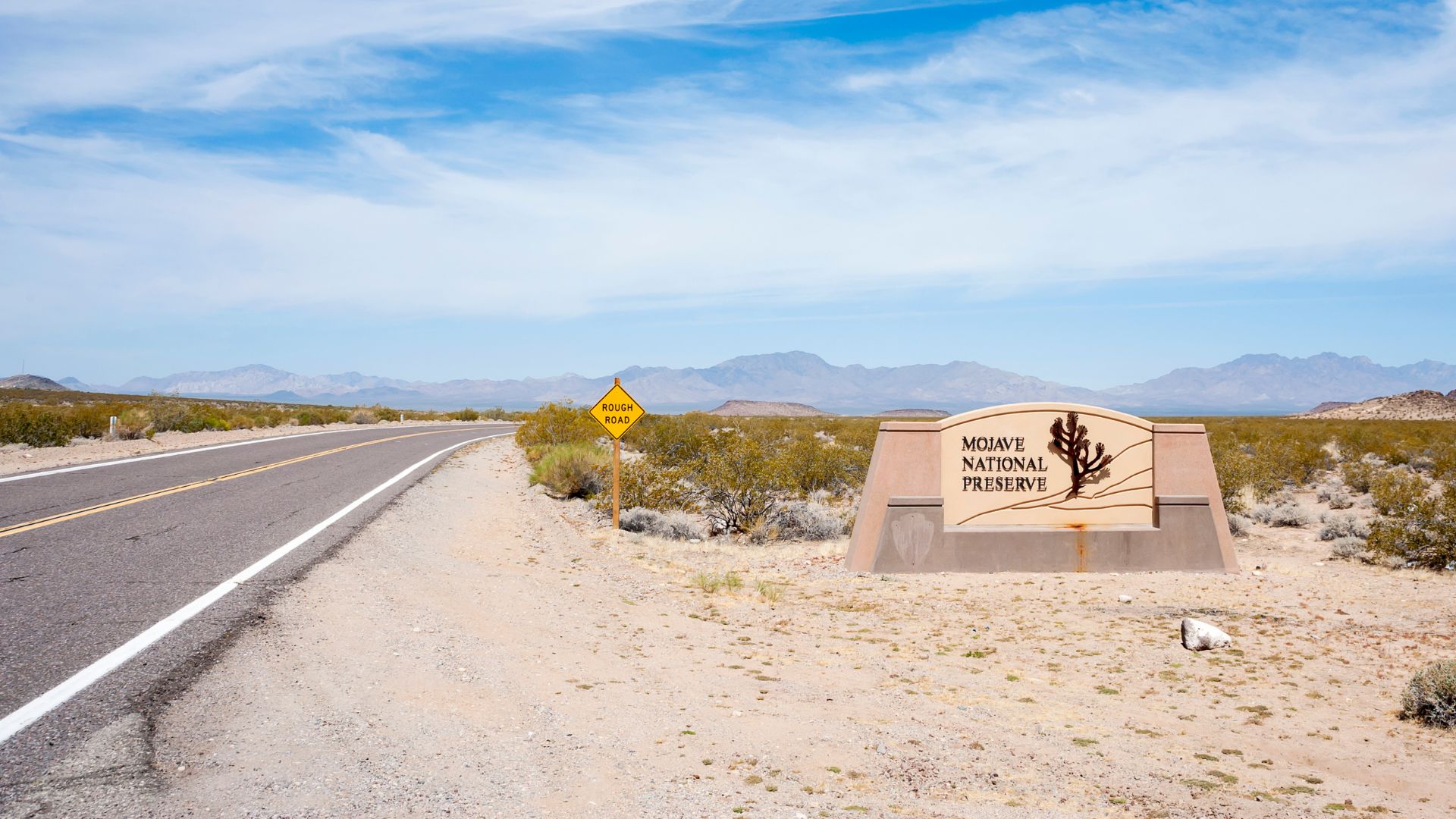 A sign for the Mojave National Preserve stands beside a desert road, with a "Rough Road" warning sign visible further down the road. The landscape features sparse desert vegetation and mountains in the background under a blue sky with scattered clouds.