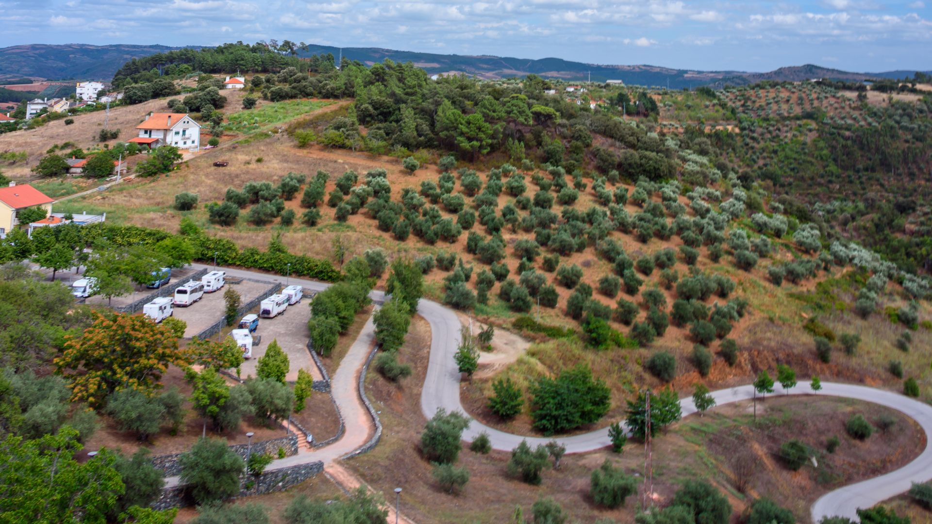 A panoramic view of a hilly, green landscape in Montesinho Natural Park, Portugal, featuring terraced olive groves, scattered houses, and a winding road descending through the terrain under a partly cloudy sky.