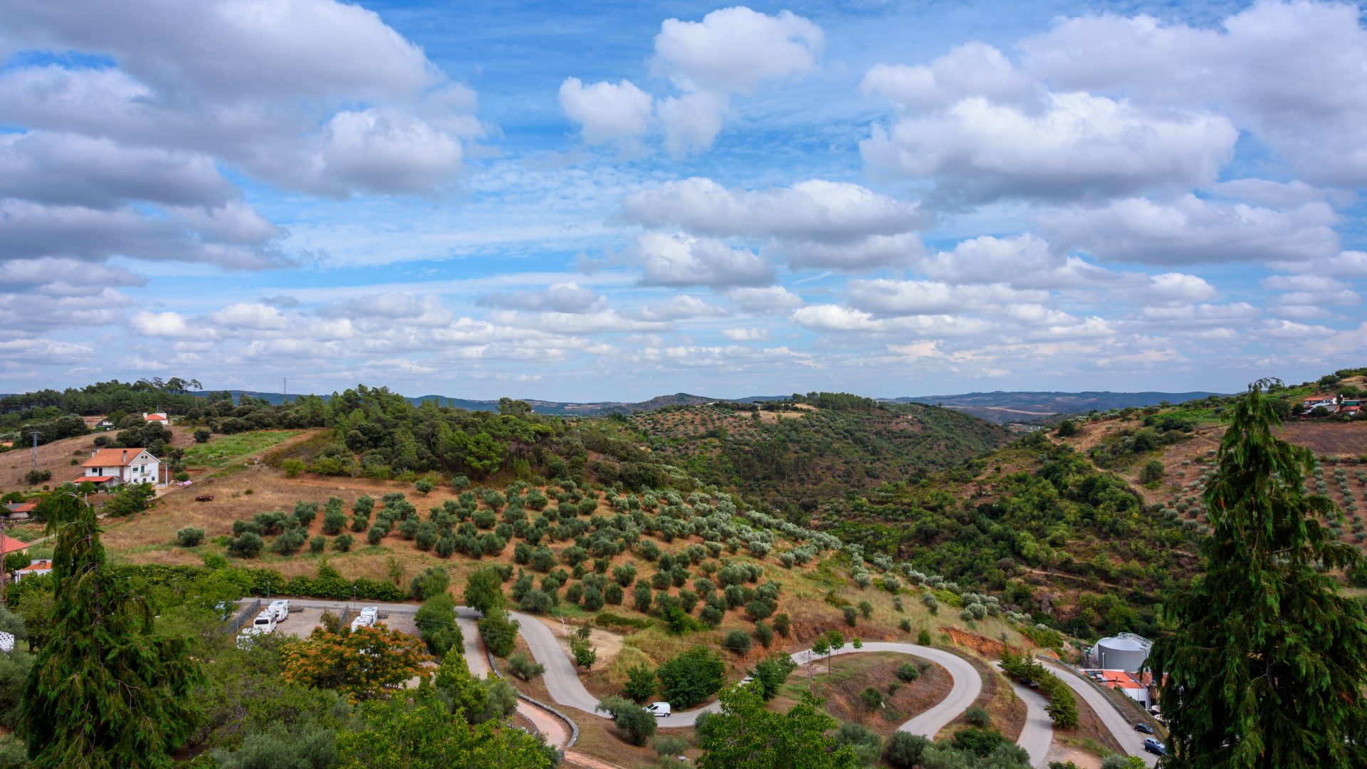 A panoramic view of a hilly, green landscape in Montesinho Natural Park, Portugal, featuring terraced olive groves, scattered houses, and a winding road descending through the terrain under a partly cloudy sky.