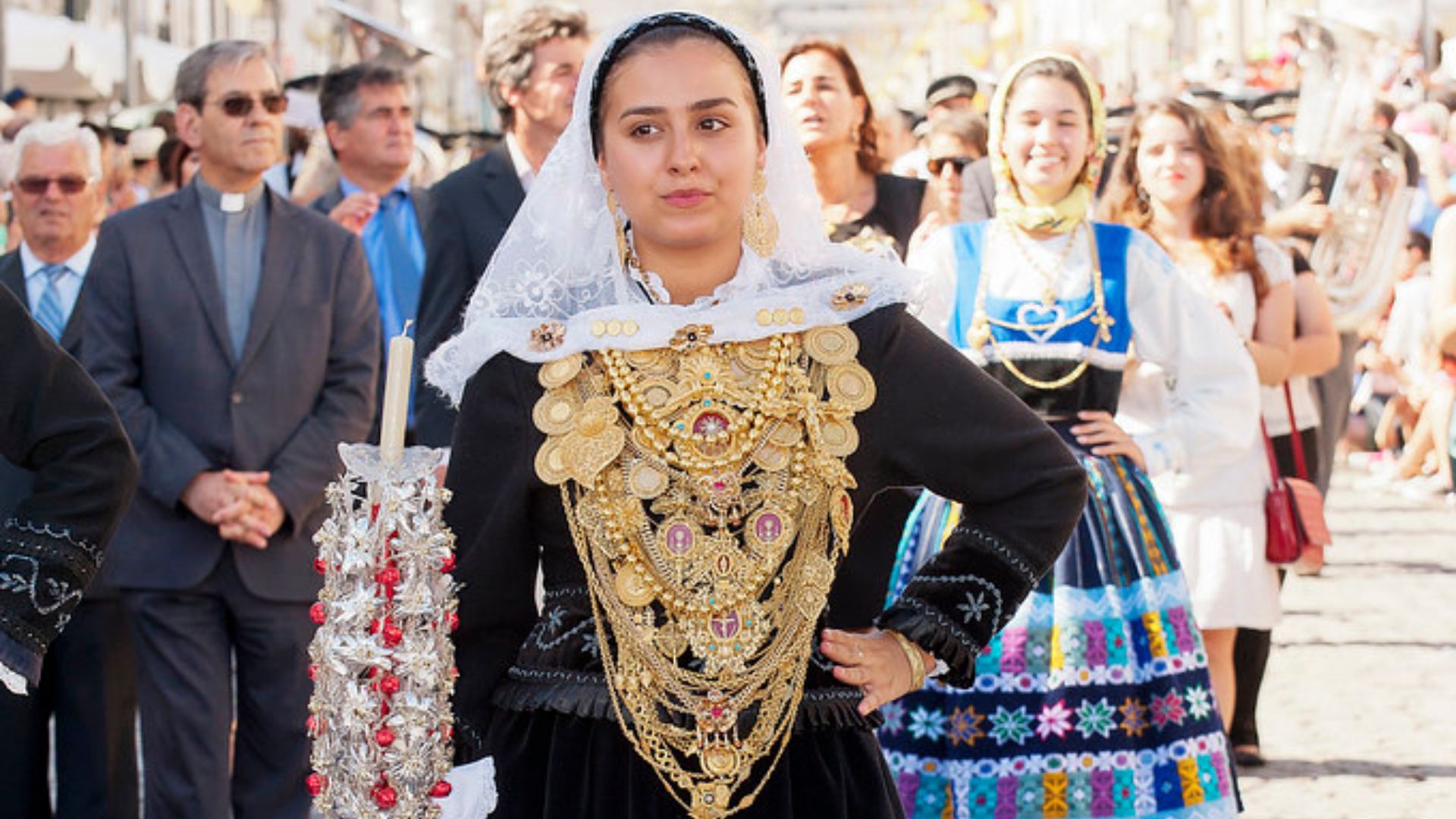A woman in a black and gold traditional Portuguese costume with ornate jewelry and a white lace head covering holds an embellished staff during a festival parade.