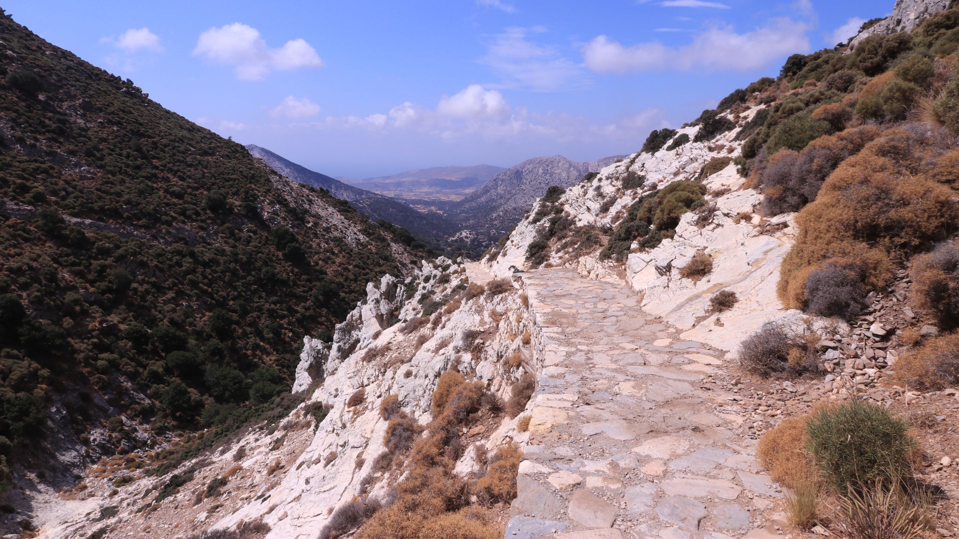 A stone path winds through a rocky mountain landscape under a blue sky with clouds.