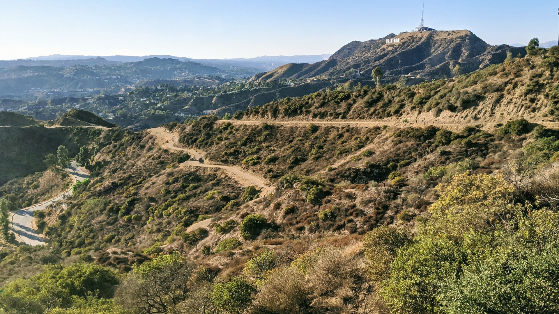 A winding dirt trail snakes up a hillside in Griffith Park, Los Angeles, with the iconic Hollywood Sign visible in the distance. The landscape is characterized by dry, scrubby vegetation under a clear blue sky. This popular hiking trail offers scenic views of the city and the Hollywood Hills.