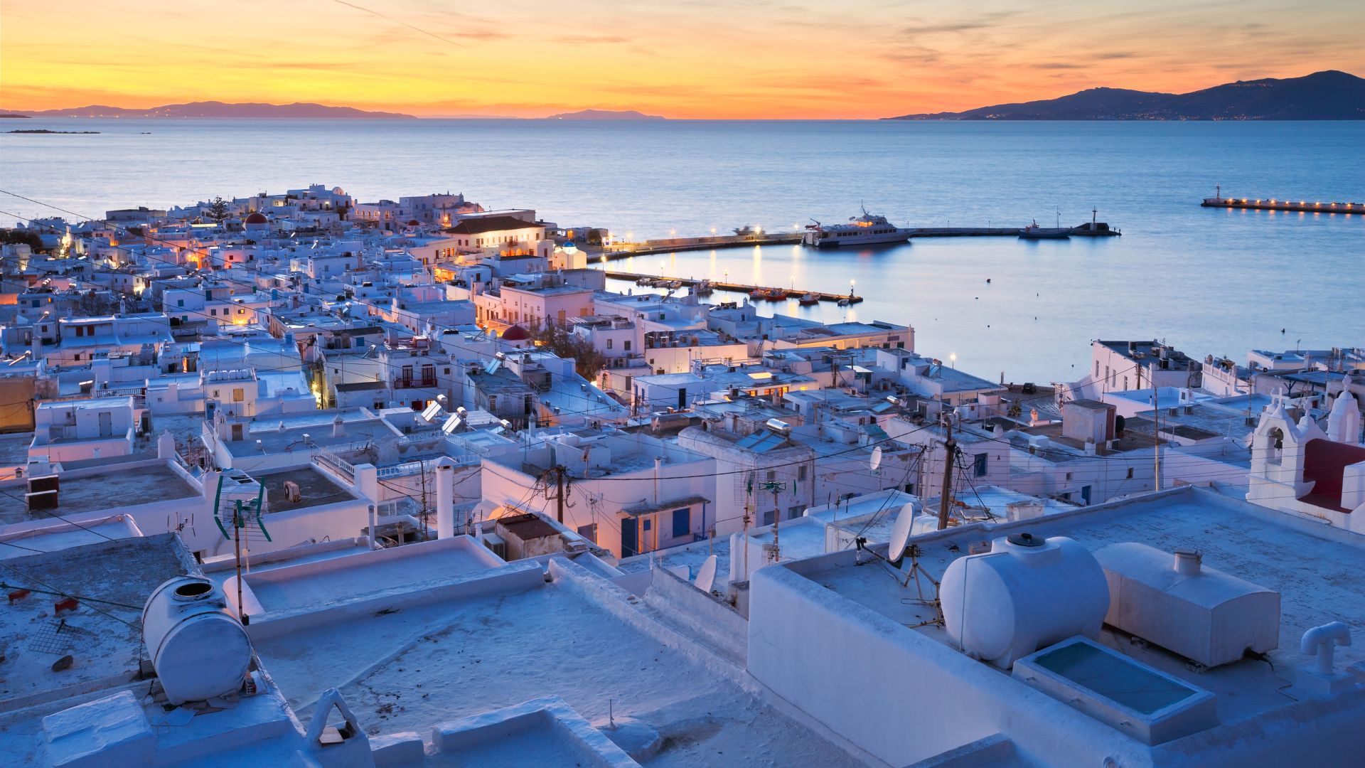 An aerial view of a Greek town with white buildings, a harbor, and a sunset sky.