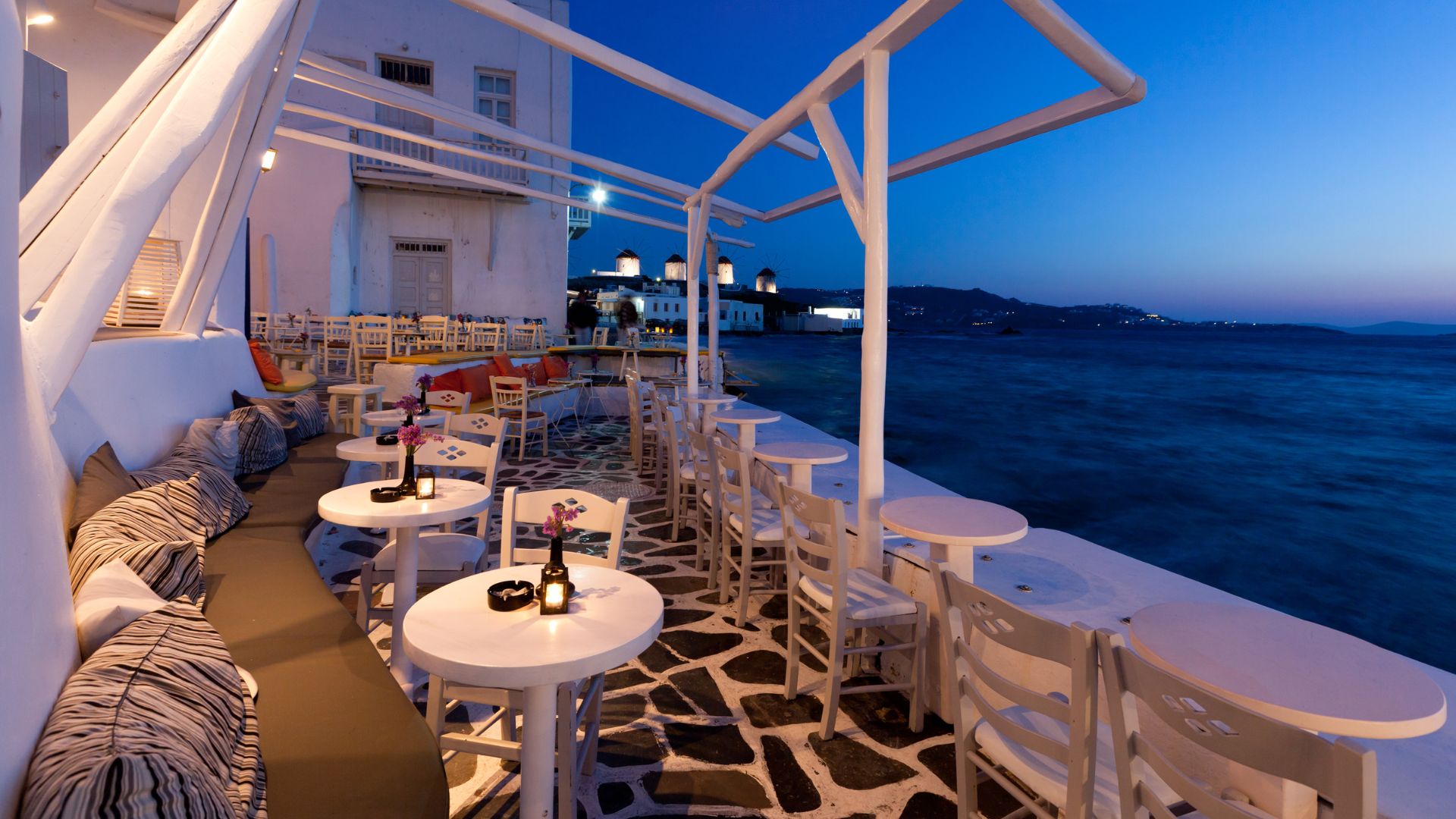 Seaside dining in Mykonos, with white tables and chairs overlooking the Aegean Sea at dusk.