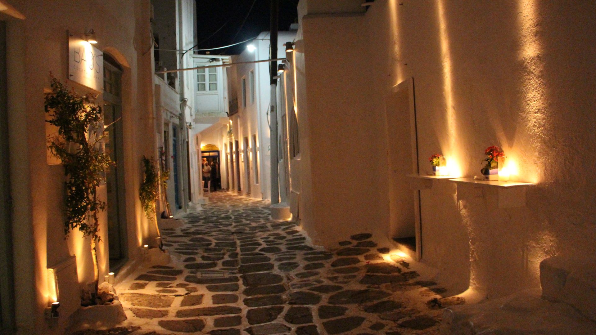 A narrow, cobblestone street in Mykonos at night, illuminated by warm lights.