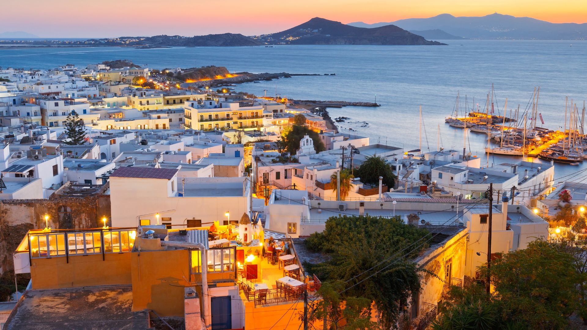 Aerial view of  Naoussa Village in Paros, Greece, featuring whitewashed buildings, mountain, sea at sunset.