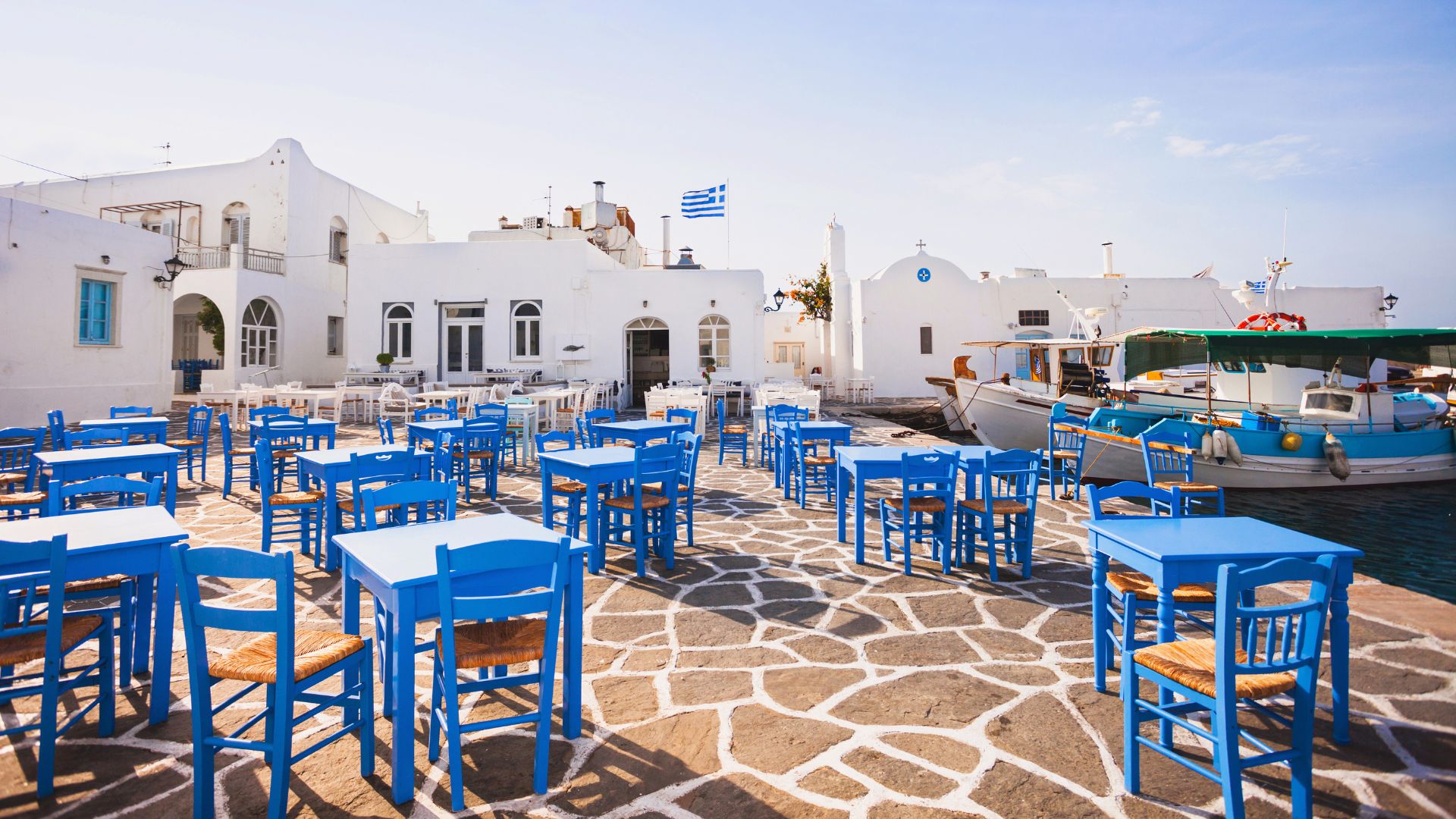 A charming village scene of a cafe in  in Naoussa Village in Paros, Greece, featuring whitewashed buildings, blue accents, and vibrant.