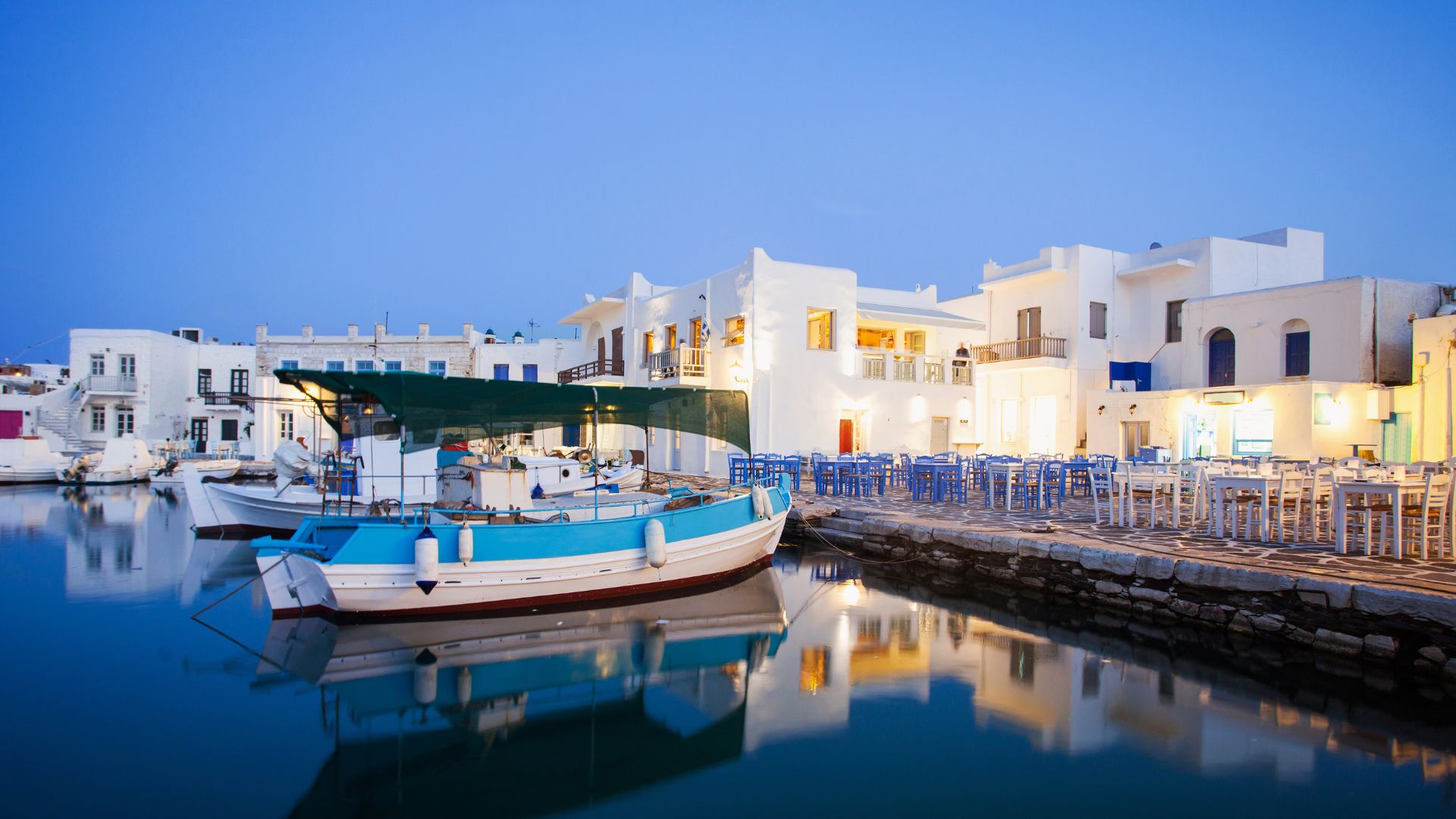 Naoussa harbor at dusk in Paros, Greece, with boats, white buildings, and blue tables.
