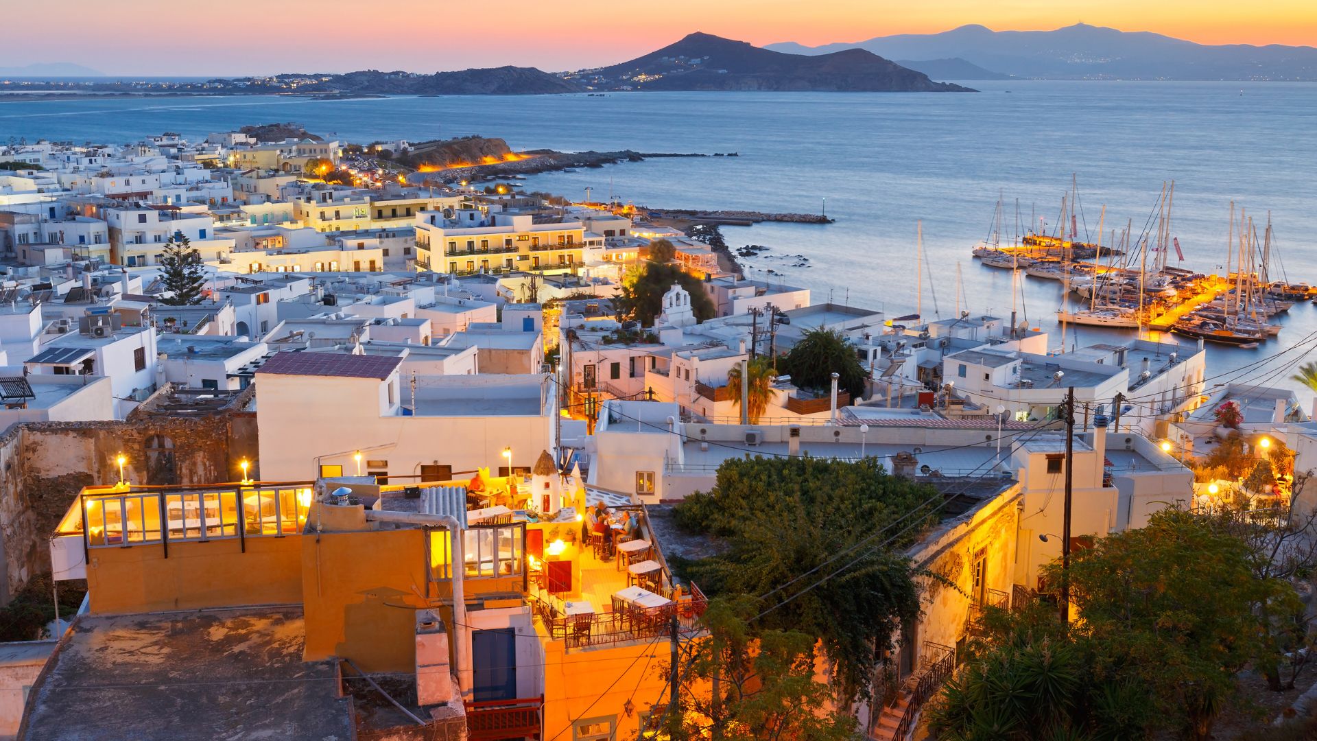 A coastal town in Greece with white buildings, a harbor with sailboats, and mountains in the background.