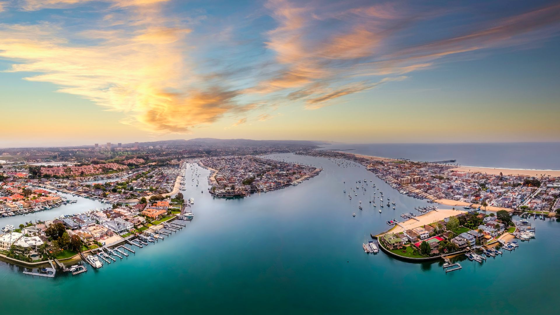 Aerial panoramic view of Newport Beach, California, featuring the stunning Newport Bay, residential areas, boats docked in the harbor, and a vibrant sunset sky over the Pacific Ocean, ideal for showcasing coastal real estate, luxury travel, and Southern California tourism.