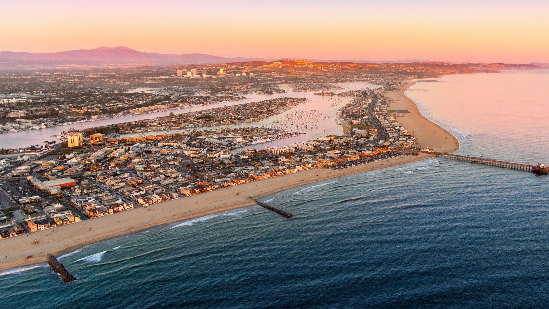 Aerial view of Newport Beach, California, at sunset, featuring the scenic coastline, yacht-filled harbor, and pier, ideal for showcasing coastal living and travel destinations