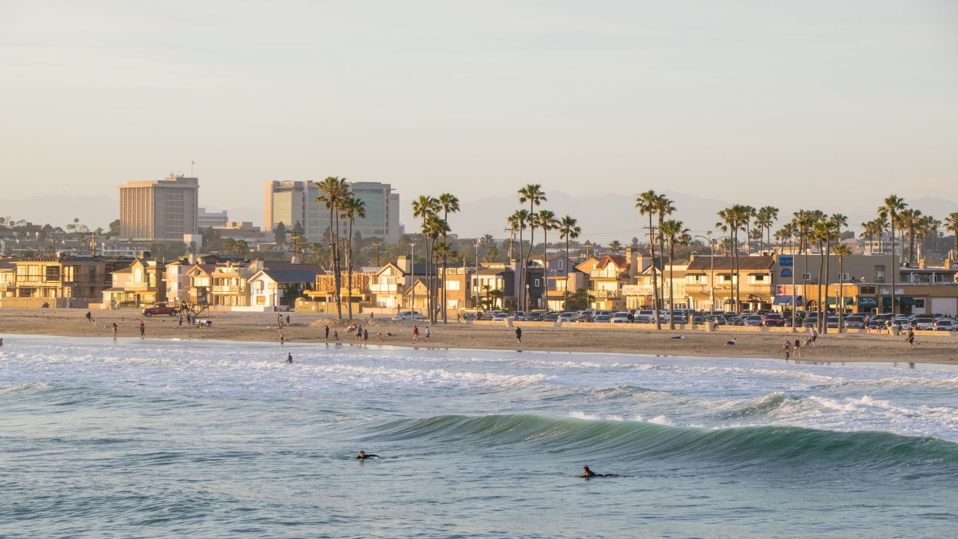 A wide shot of Newport Beach, California, at sunset, showing the ocean with gentle waves and two people on surfboards in the foreground. In the background, the sandy beach is visible with several people walking and buildings with palm trees lining the coastline. 