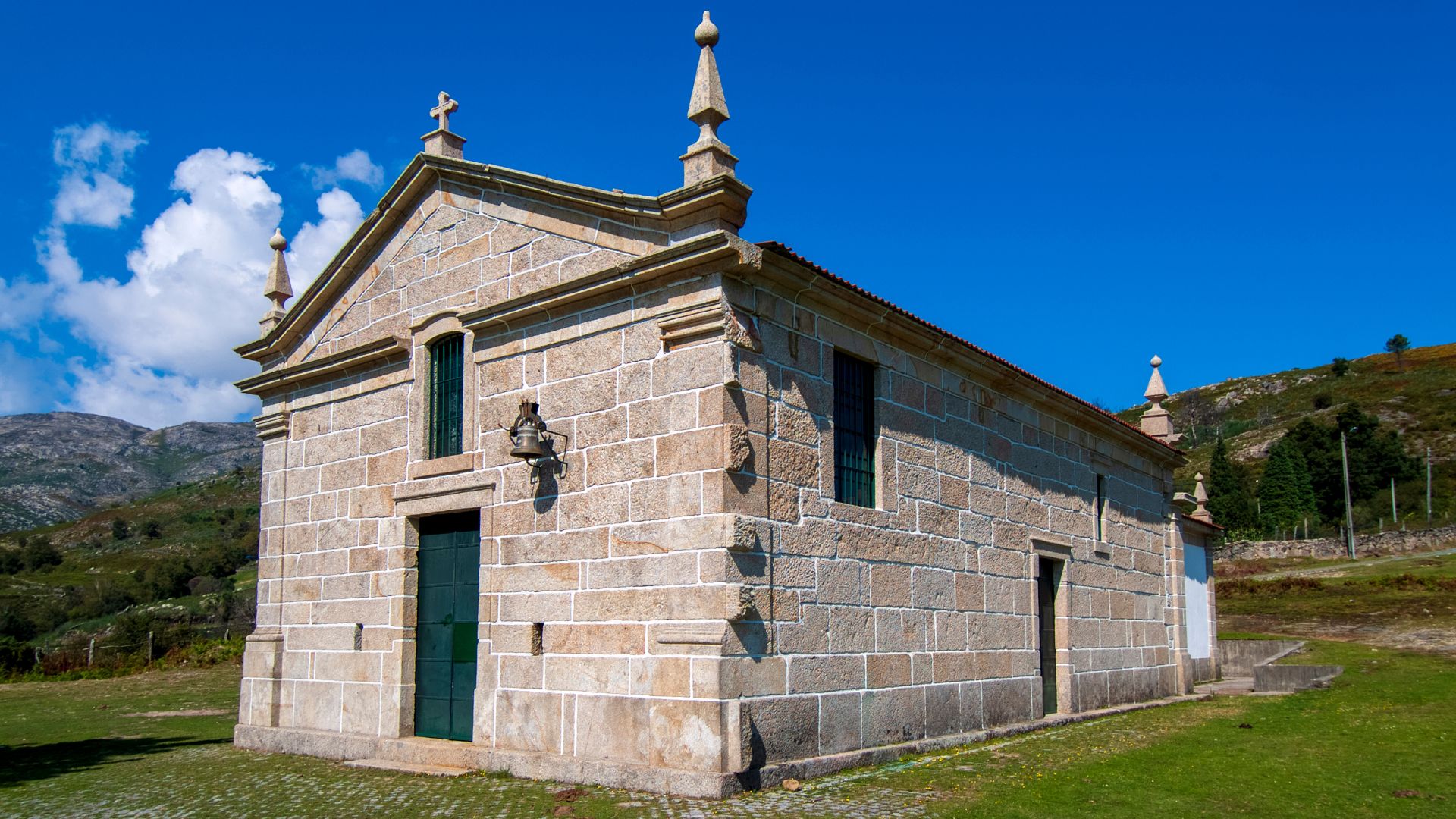 A stone chapel with a pitched roof and a small bell tower, set against a bright blue sky and green hills in Portugal.