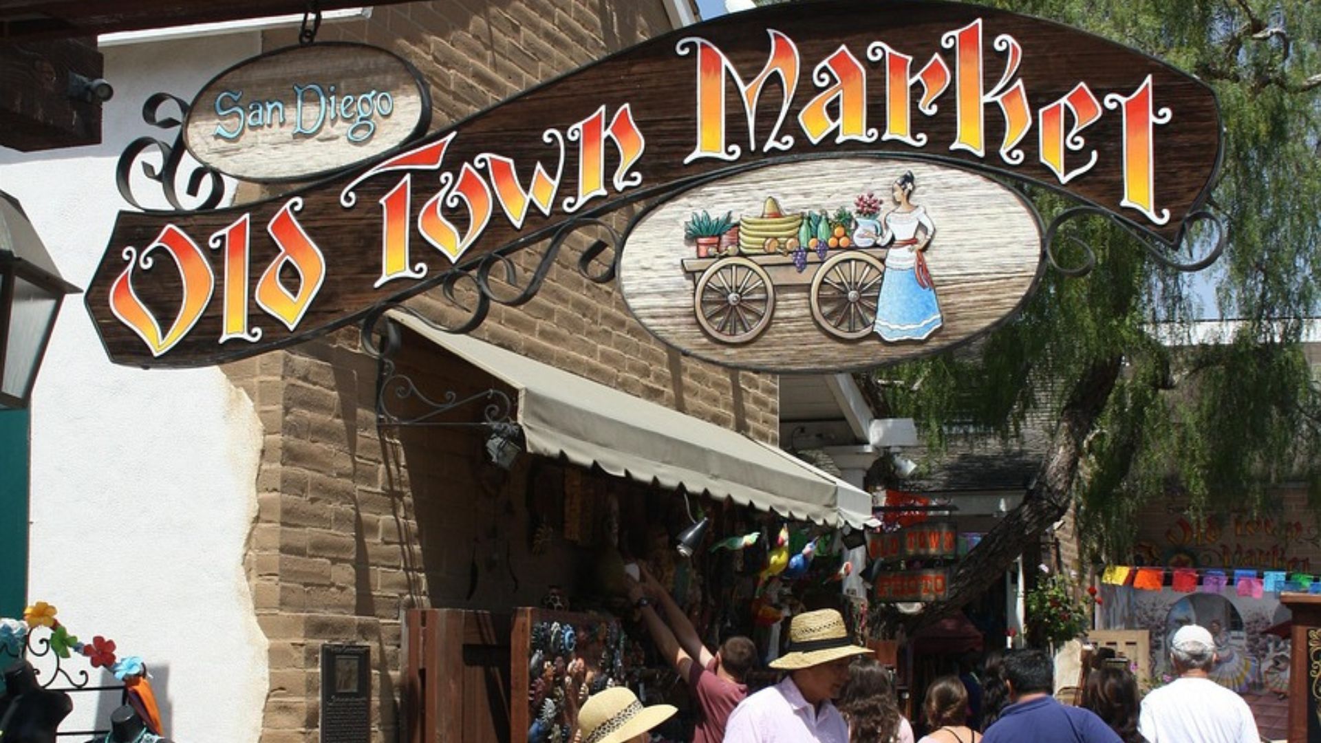 A bright, sunny outdoor shot showing the entrance to the Old Town Market in San Diego, California, with a prominent sign overhead and people walking on a paved pathway.