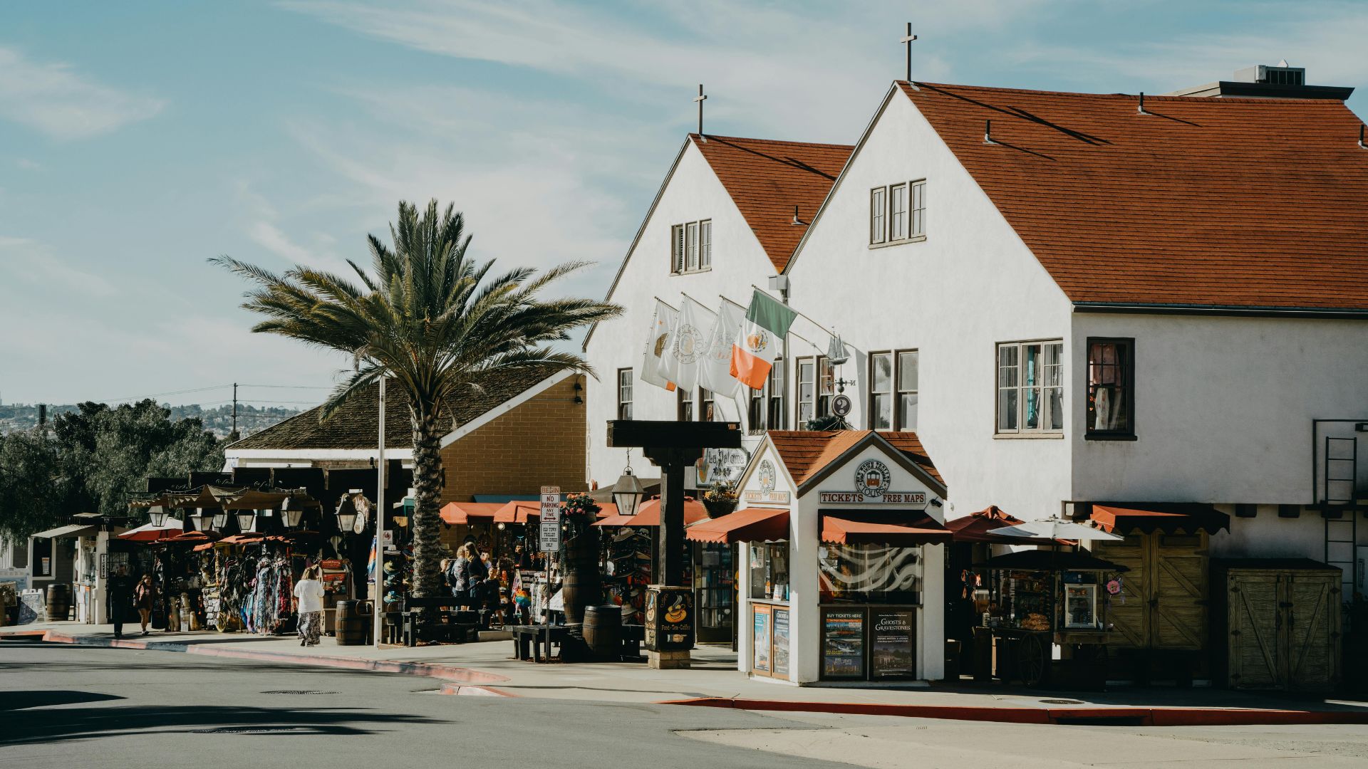 A street scene in Old Town San Diego, California, featuring historic white buildings with red tile roofs, a palm tree, and various shops and market stalls lining the street, with people browsing and walking.