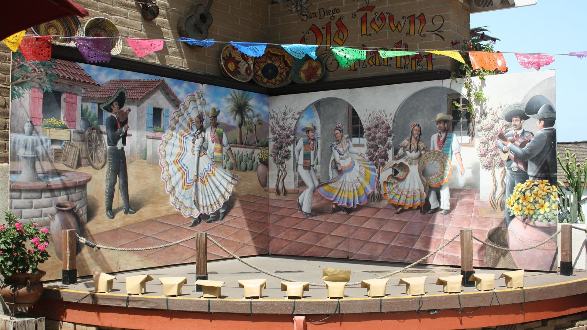 A vibrant mural in Old Town San Diego, California, depicting various figures in traditional Mexican attire dancing and celebrating in a lively outdoor setting, with colorful papel picado banners strung overhead and a stage in the foreground.