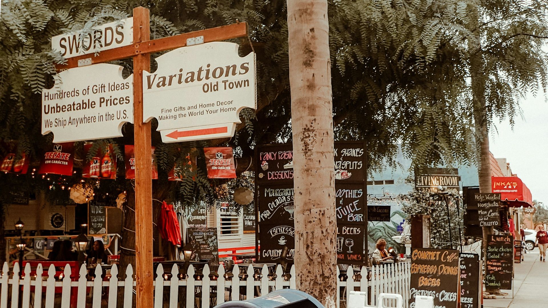 A street scene in Old Town, San Diego, featuring a blue US mailbox covered in stickers and graffiti in the foreground, with various shops and businesses with signs like "SWORDS" and "Variations Old Town" and a "GOING OUT FOR YOUR BUSINESS 40% Off" sign in the background.