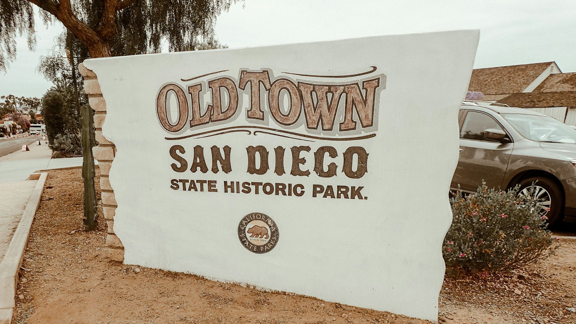 A large, white sign with brown lettering reads "OLD TOWN SAN DIEGO STATE HISTORIC PARK." Below the text is a circular emblem with a bear, indicating it is a California State Park. The sign stands on a dirt patch with some greenery, and a car and building are visible in the background. 