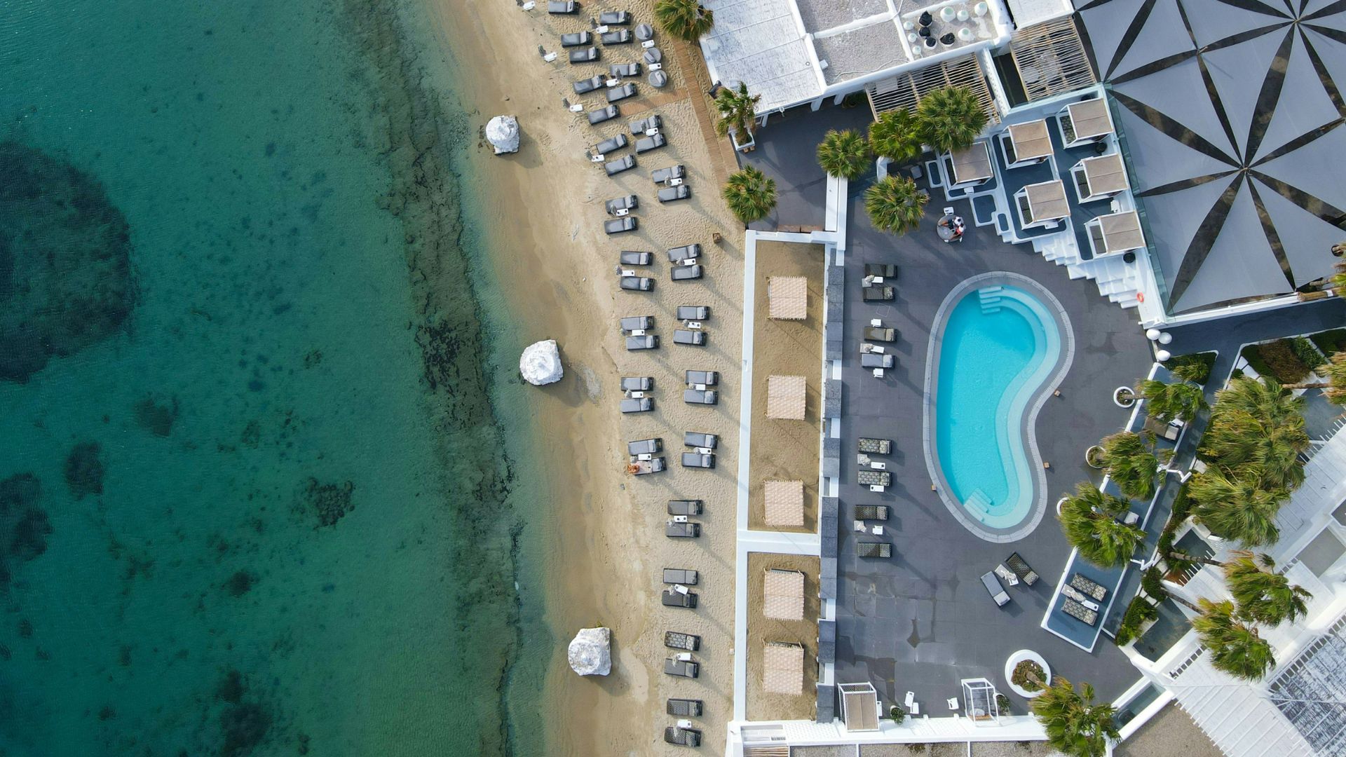 Aerial view of Ornos Beach in Mykonos, Greece, showing a resort with a pool, beach chairs, and clear turquoise water.