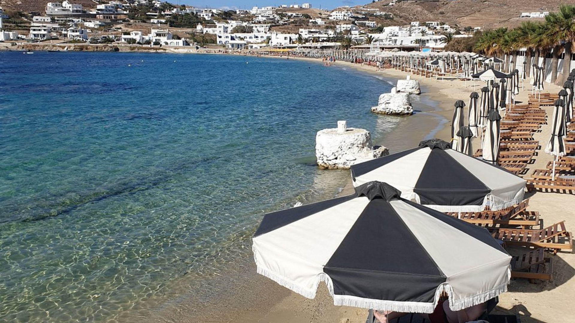 A sunny day at Ornos Beach in Mykonos, featuring calm waters and white buildings on the hills.