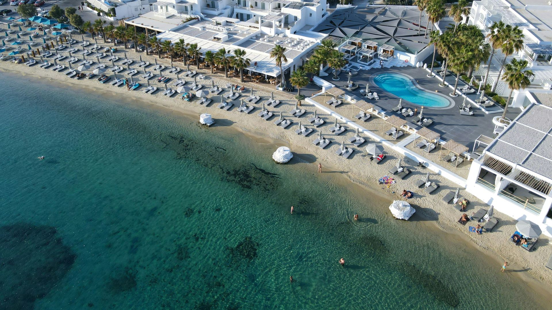 Aerial view of Ornos Beach in Mykonos, Greece, showing a resort with a pool, beach chairs, and clear turquoise water.