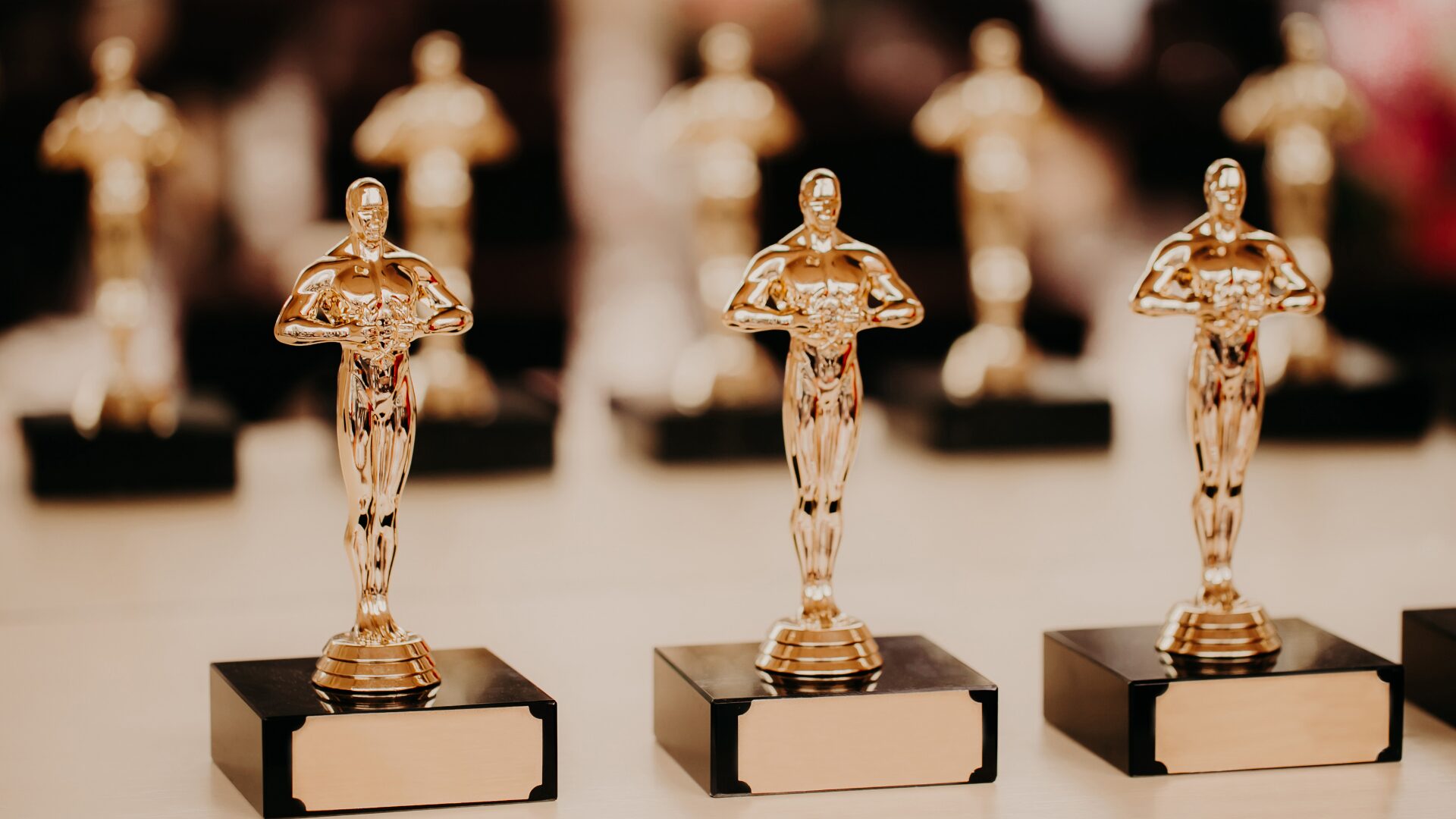 Multiple golden Oscar statuettes lined up on a light-colored surface, with a blurred background showing more awards. The image showcases the iconic design of the Academy Awards, highlighting the prestige and recognition associated with the ceremony.