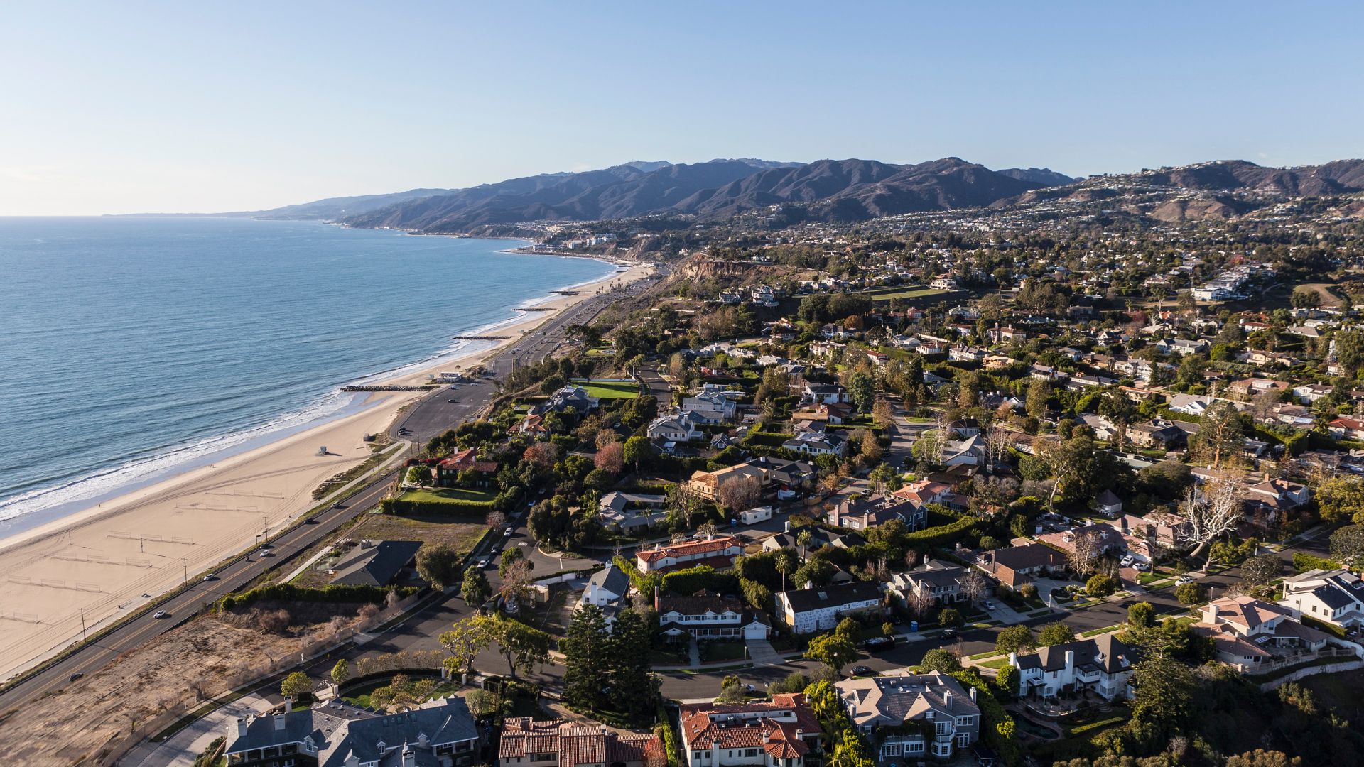 Aerial view of the Pacific Coast Highway running along a sandy beach and the Pacific Ocean, with residential areas and the Santa Monica Mountains rising in the background under a clear sky in Pacific Palisades, Los Angeles.