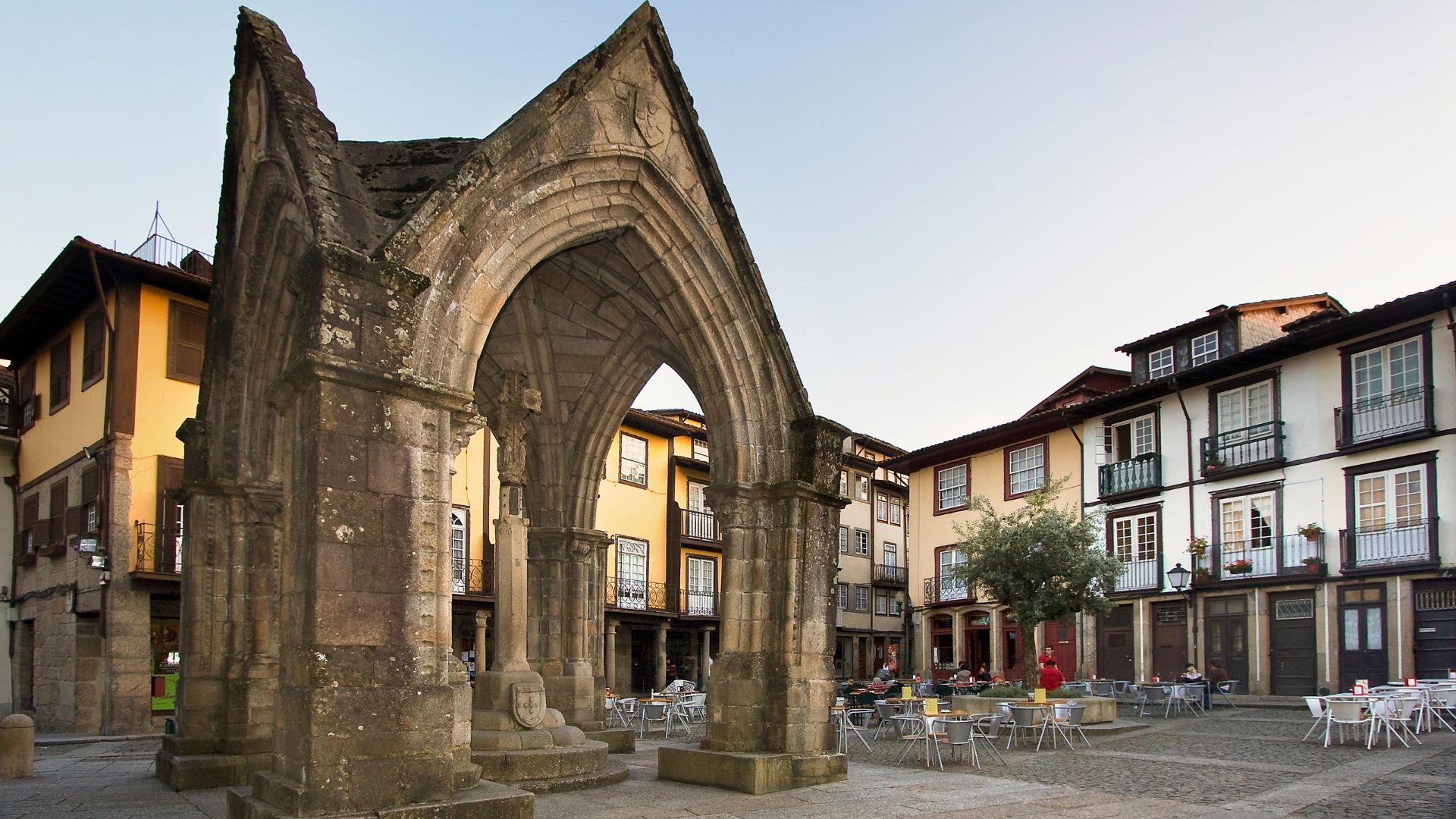 A historic stone archway, the Padrão do Salado, stands in a cobblestone square in Guimarães, Portugal, surrounded by traditional buildings and outdoor cafe seating.