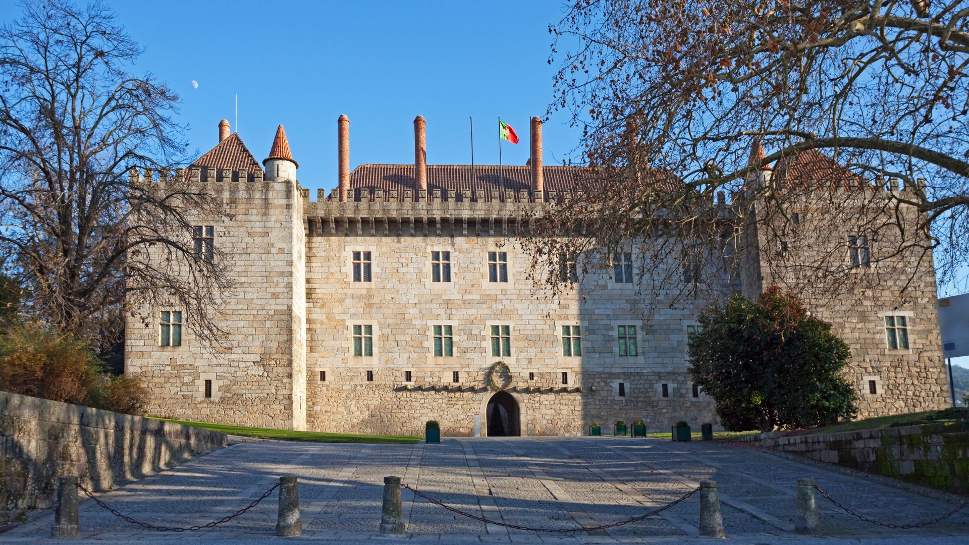 A large, historic stone palace with a prominent entrance and multiple windows, surrounded by trees and a paved area, under a clear blue sky.
