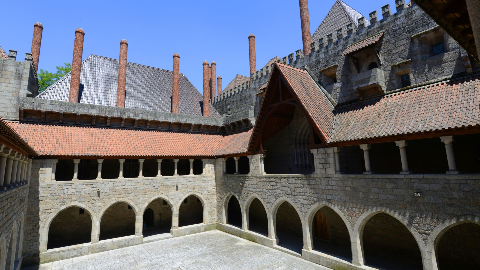 A view of the courtyard within the Palace of the Dukes of Braganza in Guimarães, Portugal, featuring stone arches, tiled roofs, and prominent chimneys.