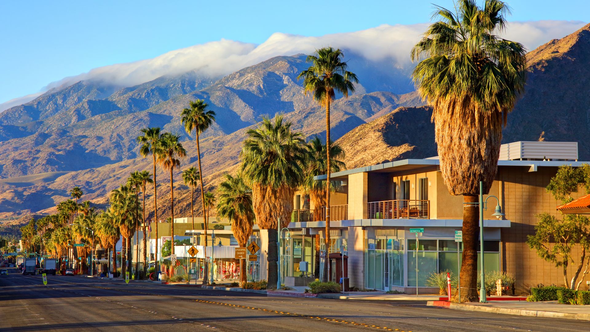A street in Palm Springs, California, lined with tall palm trees and buildingas, with mountains in the background under a clear sky with some clouds. 