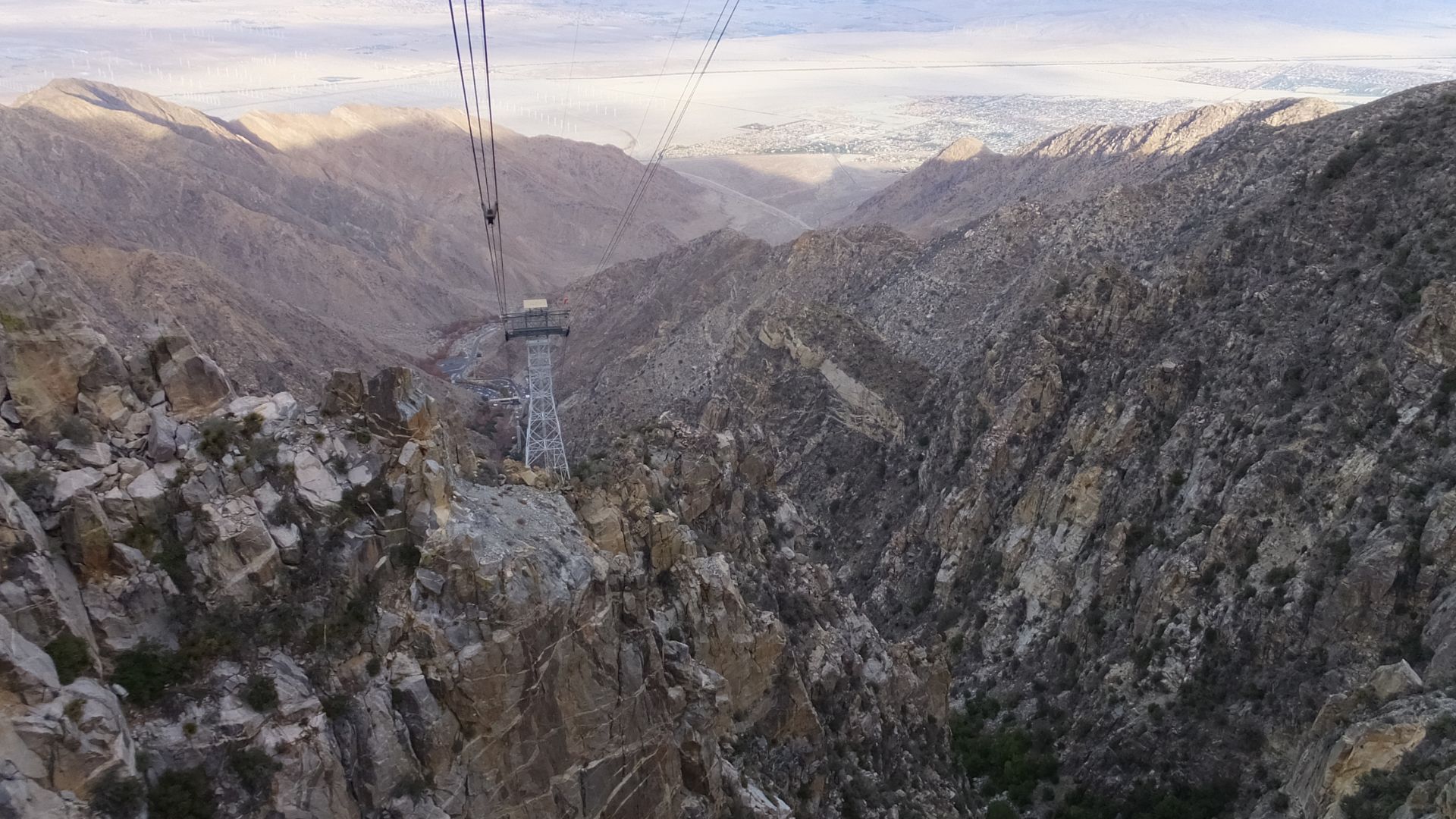 A view from the Palm Springs Aerial Tramway looking down into Chino Canyon, showing a tall metal tramway tower in the foreground and the desert valley and city in the distance under a clear sky.