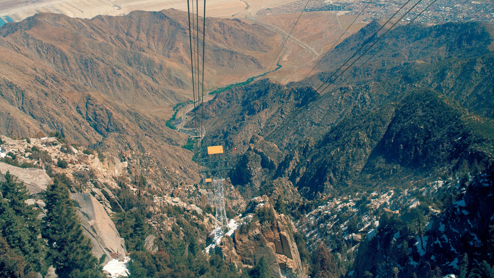 A view from the Palm Springs Aerial Tramway looking down into Chino Canyon, showing a tall metal tramway tower in the foreground and the desert valley and city in the distance under a clear sky.