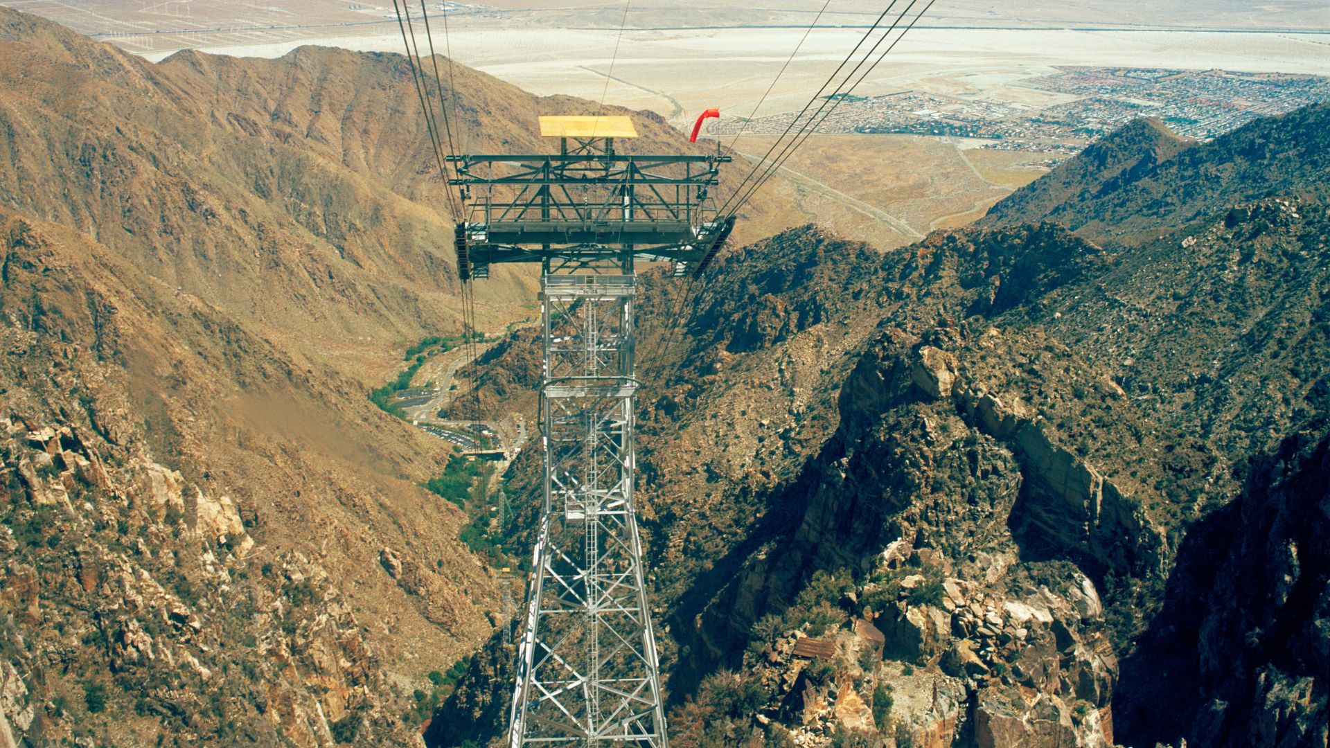 A view from the Palm Springs Aerial Tramway looking down into Chino Canyon, showing a tall metal tramway tower in the foreground and the desert valley and city in the distance under a clear sky.