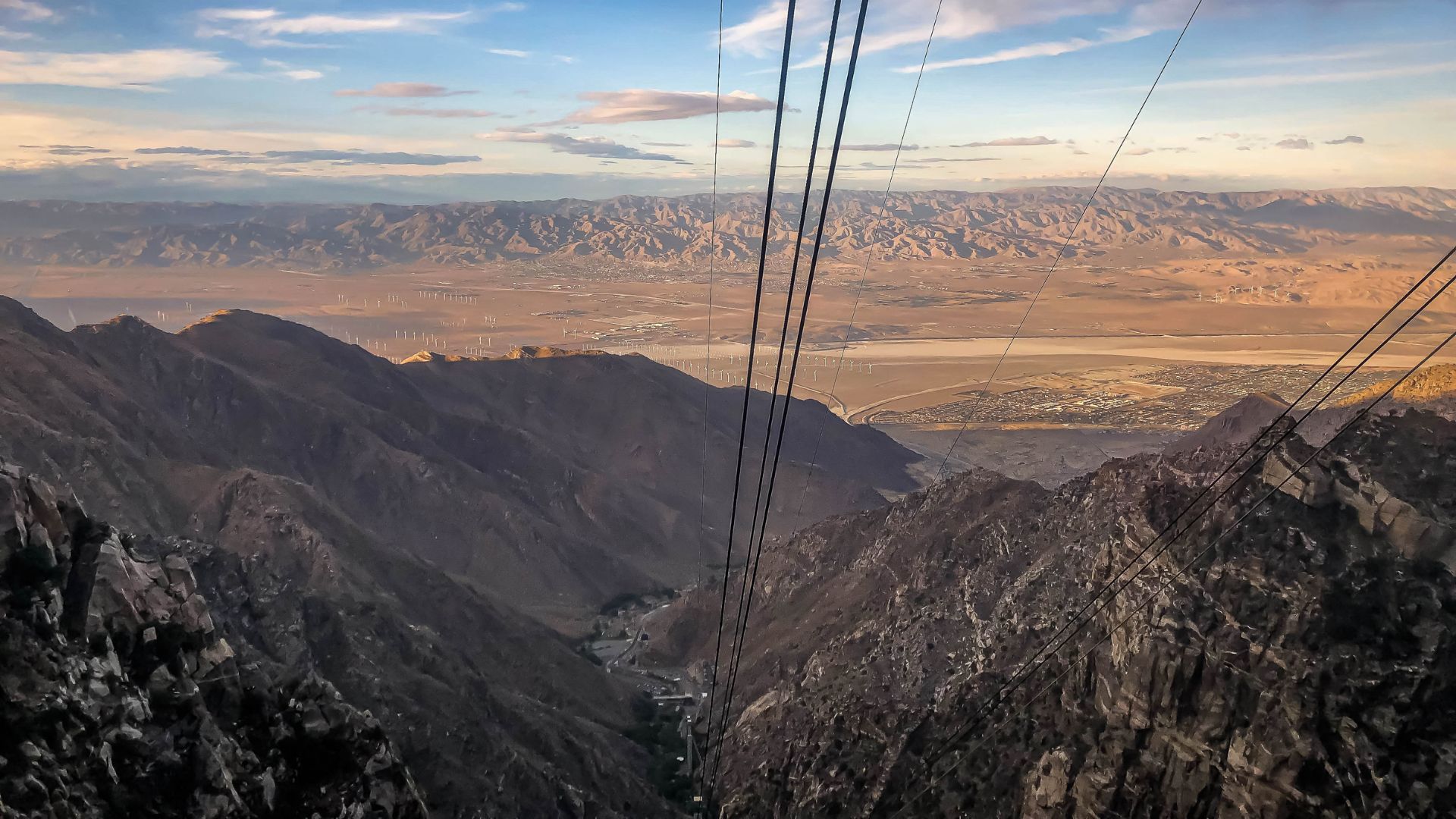 A view from a tram car looking down into a vast canyon with rocky mountains on either side, and a distant desert valley under a partly cloudy sky. The cables of the Palm Springs Aerial Tramway are visible in the foreground.