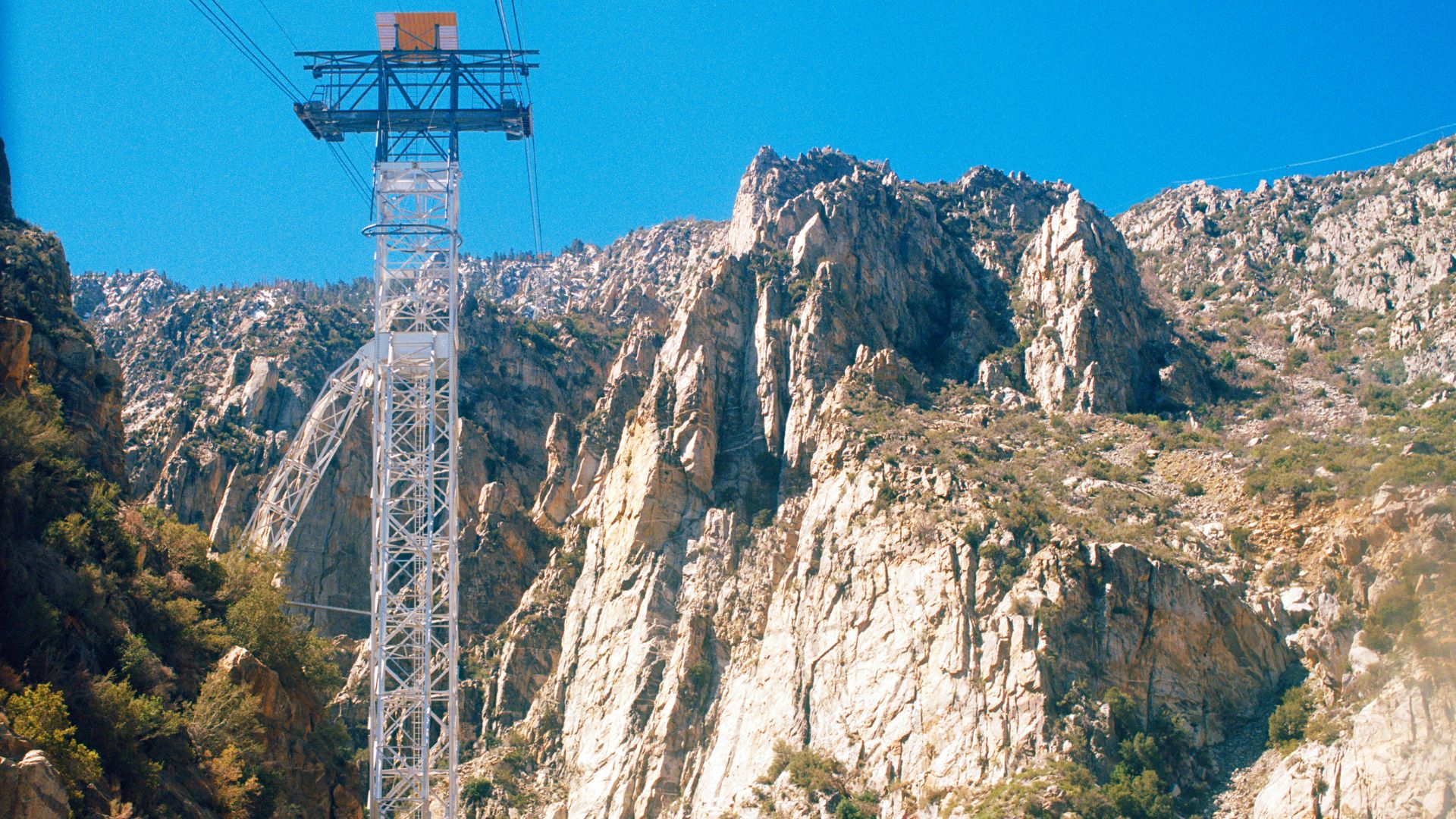 A view from the Palm Springs Aerial Tramway looking down into Chino Canyon, showing a tall metal tramway tower in the foreground and the desert valley and city in the distance under a clear sky.