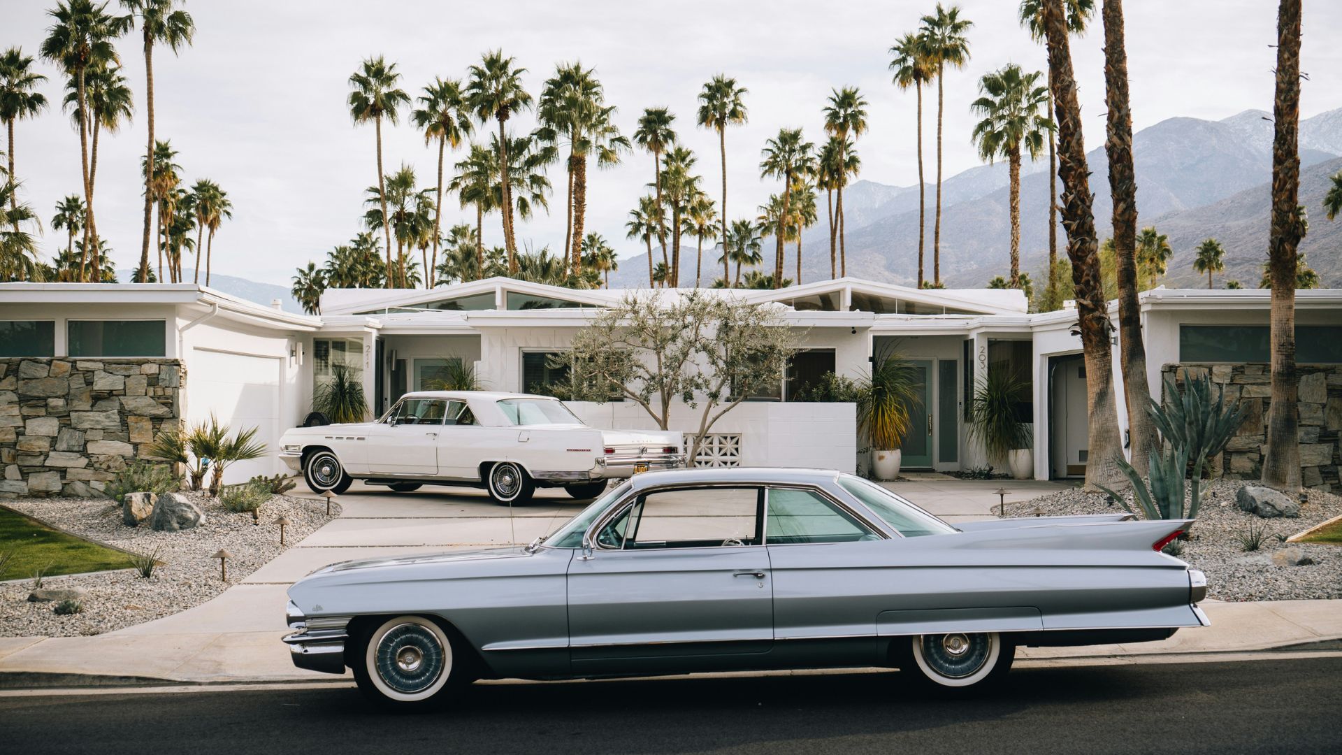 A classic silver 1961 Cadillac Coupe de Ville is parked on a street in front of a white mid-century modern house with a stone facade, surrounded by tall palm trees and desert landscaping under a bright sky in Palm Springs, California. Another vintage white car is visible in the driveway of the house.