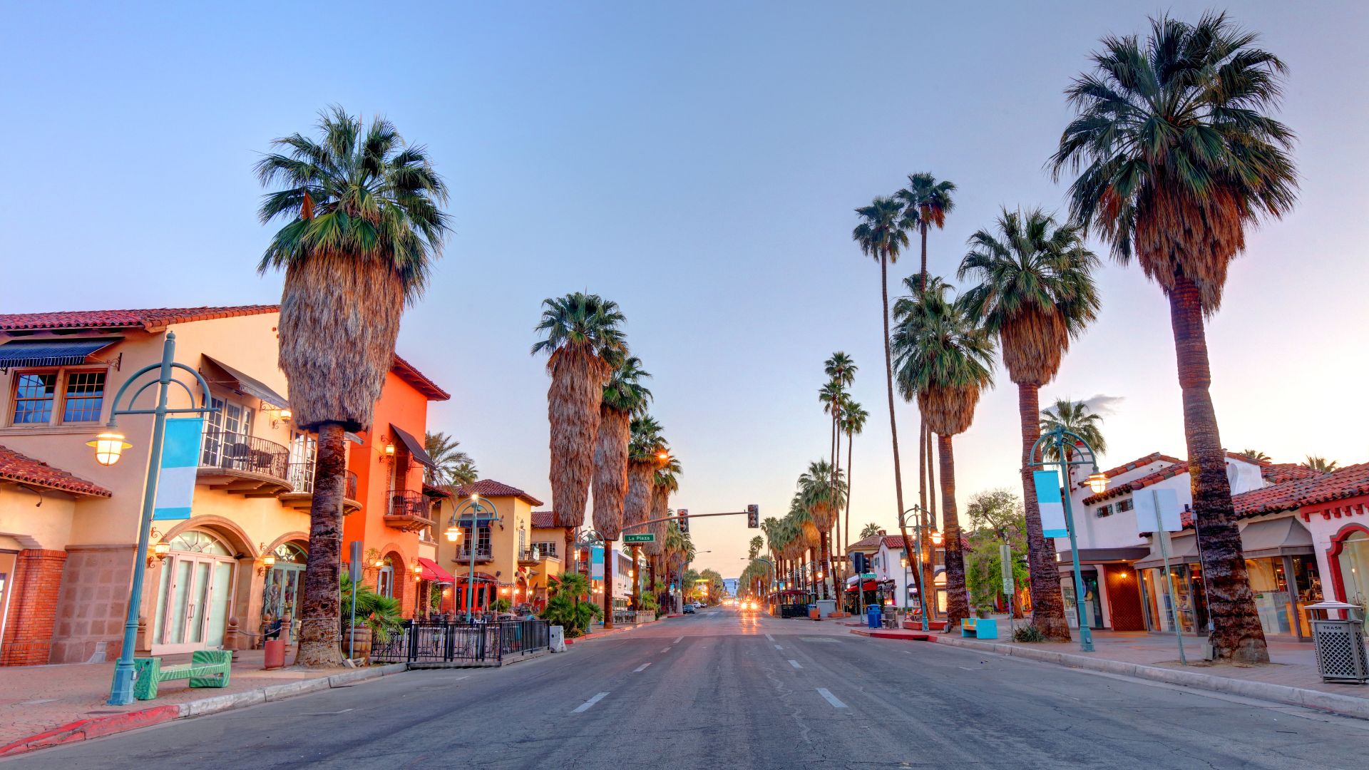 Palm Springs street scene at dusk. Palm trees line the road with historic buildings in the background. The sky is a gradient of blue and pink.