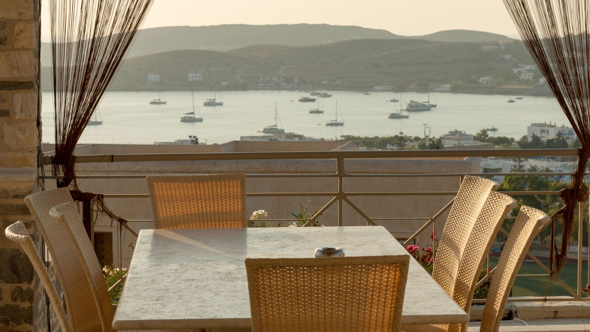 A cafe on a terrace at Paros, Greece overlooking the boats, mountains, white building, and the sea.