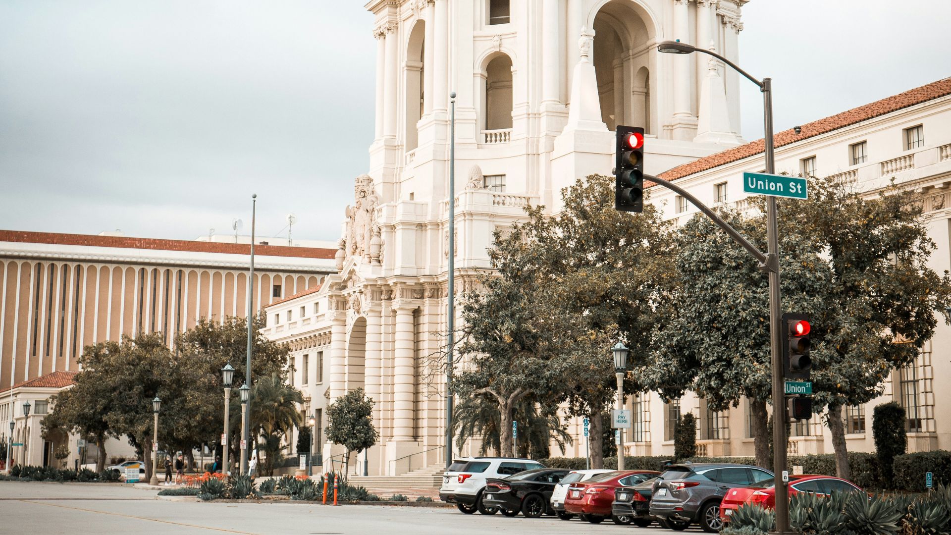 A grand, light-colored building with a prominent domed roof and classical architecture, identified as Pasadena City Hall, stands under an overcast sky with a street and parked cars in the foreground. A street sign for "Union St" and traffic lights are visible.