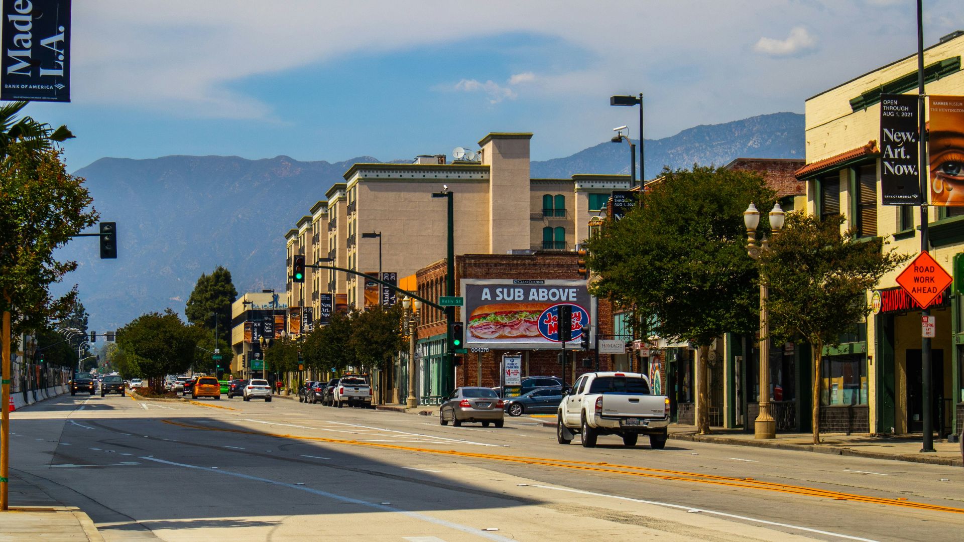 A daytime street scene in Pasadena, California, showing a tree-lined street with cars, buildings with storefronts, and mountains in the background under a partly cloudy sky. One prominent sign for "A SUB ABOVE" is visible on a building.