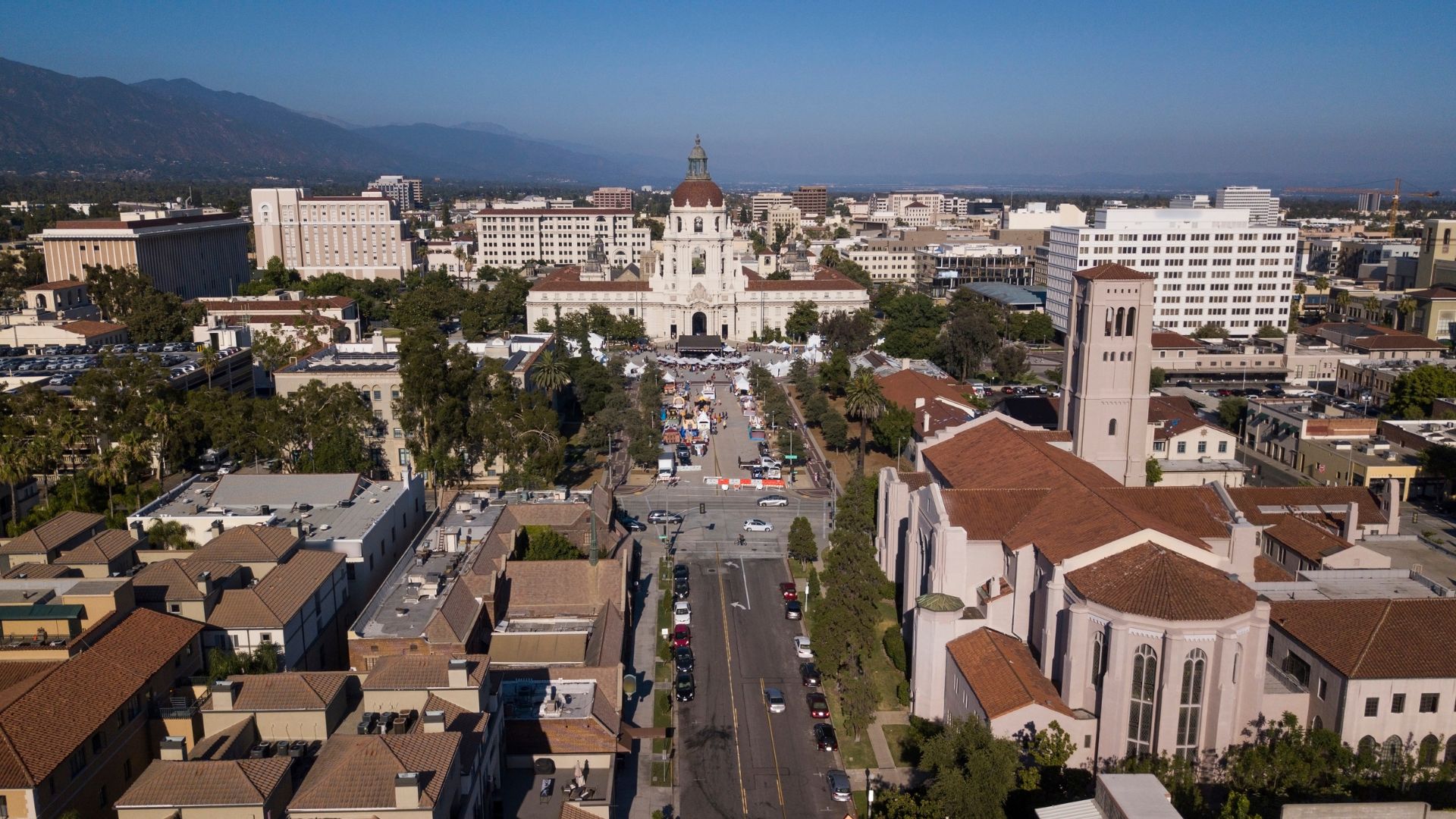 An aerial view of Pasadena, California, showing the iconic Pasadena City Hall and surrounding buildings under a clear sky, with mountains visible in the background. 