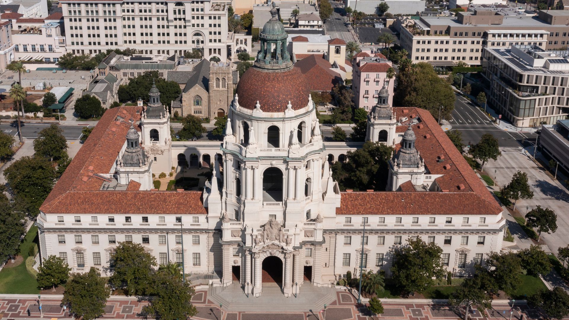 An aerial view of Pasadena City Hall, a large, ornate building with a central dome and surrounding wings, situated within a city landscape.