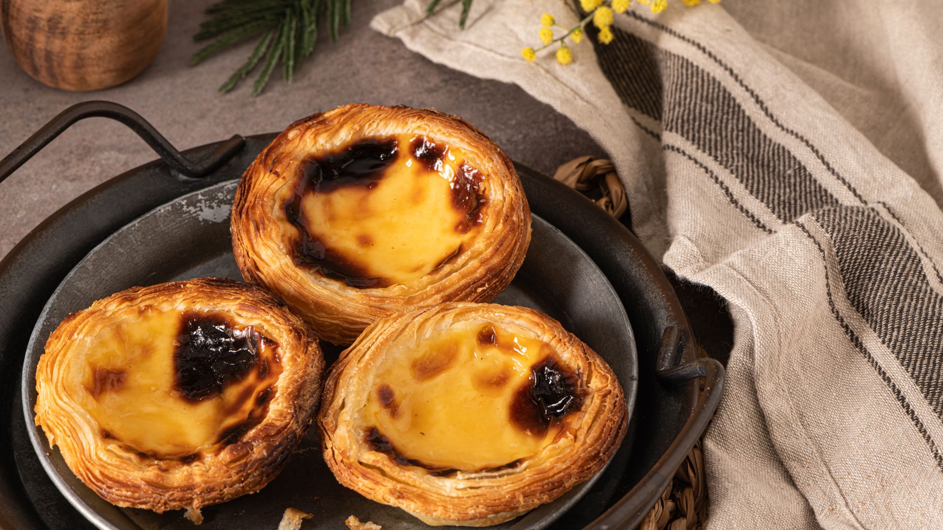 A close-up shot of three pastel de nata arranged on a metal plate, with a linen cloth in the background. The tarts have a golden-brown, flaky crust and a creamy, slightly burnt custard filling.
