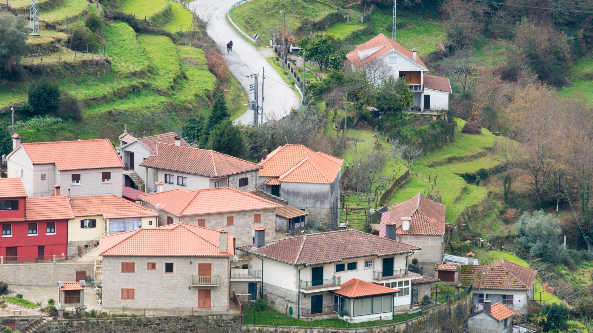 A village with red-tiled roofs nestled in a green, terraced mountain landscape with a winding road.