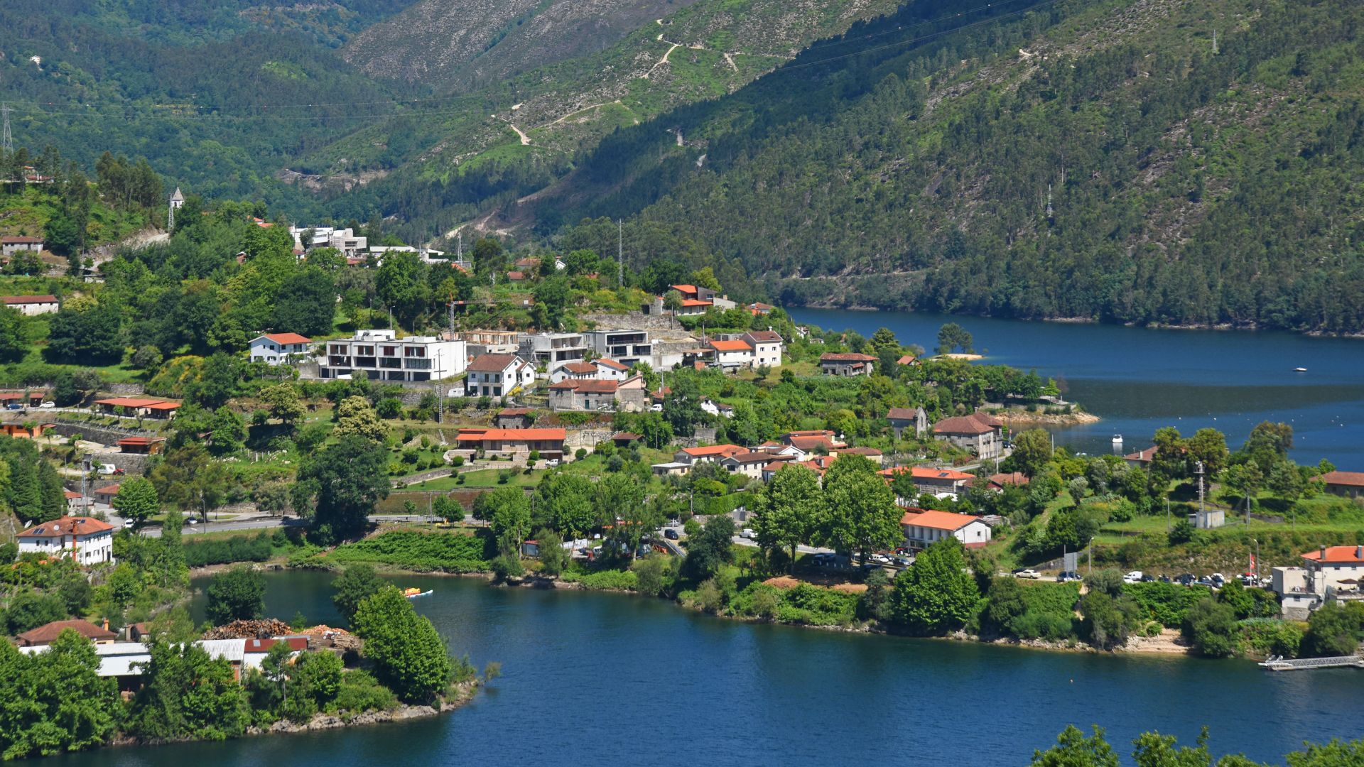 An aerial view of a village nestled by a lake within a mountainous, forested landscape under a clear sky.